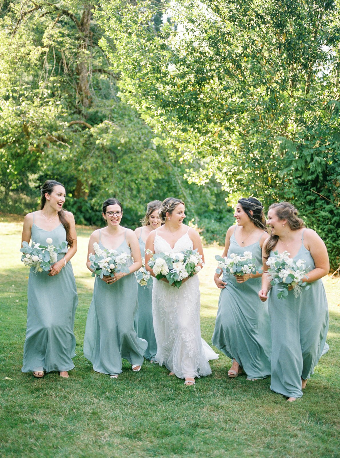 Bridesmaids in sage blue dresses walking through Abigail’s Garden with white and eucalyptus bouquets.