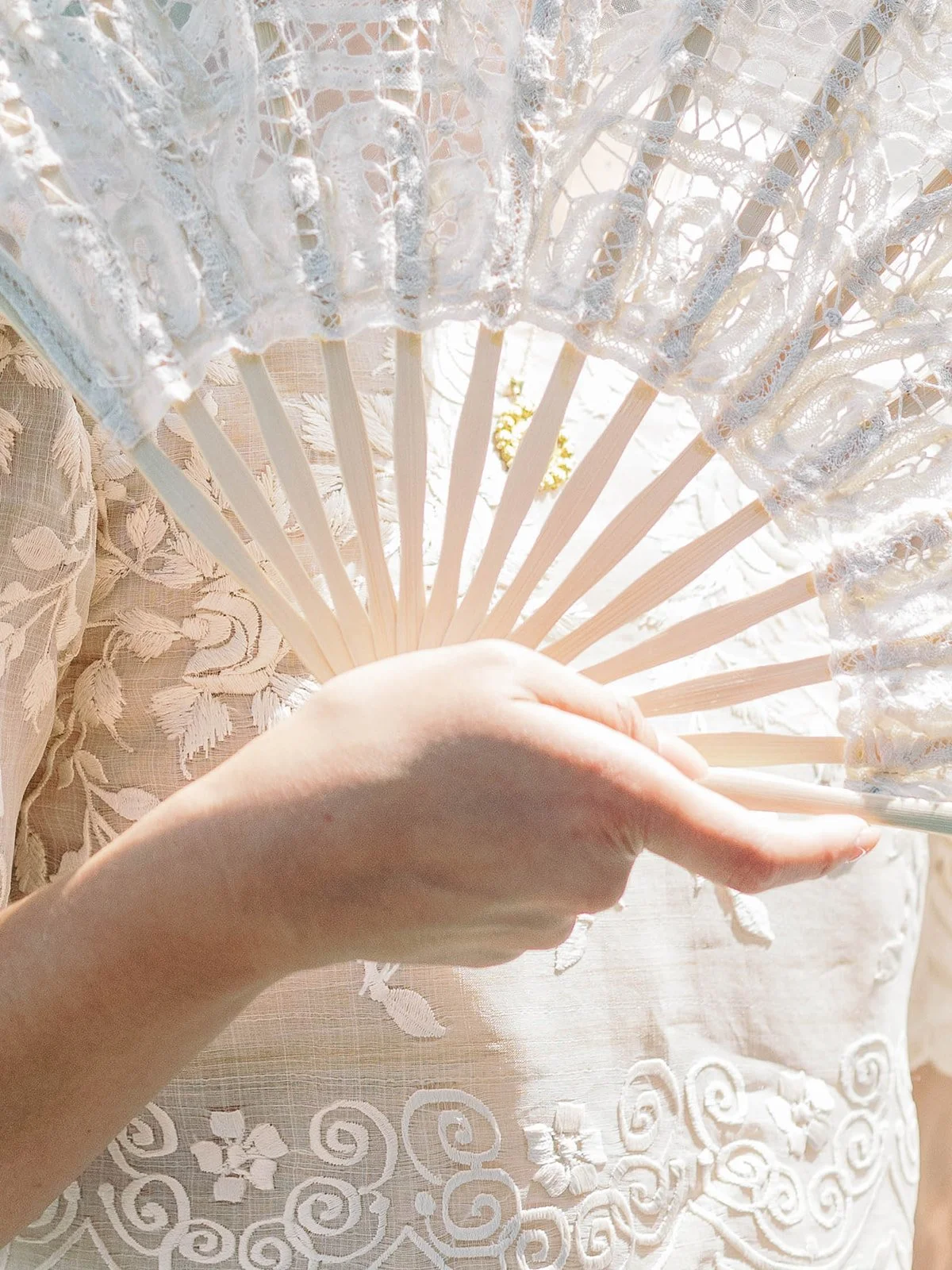 Close-up of a bride's hand holding an open white lace fan against her embroidered Filipino wedding gown
