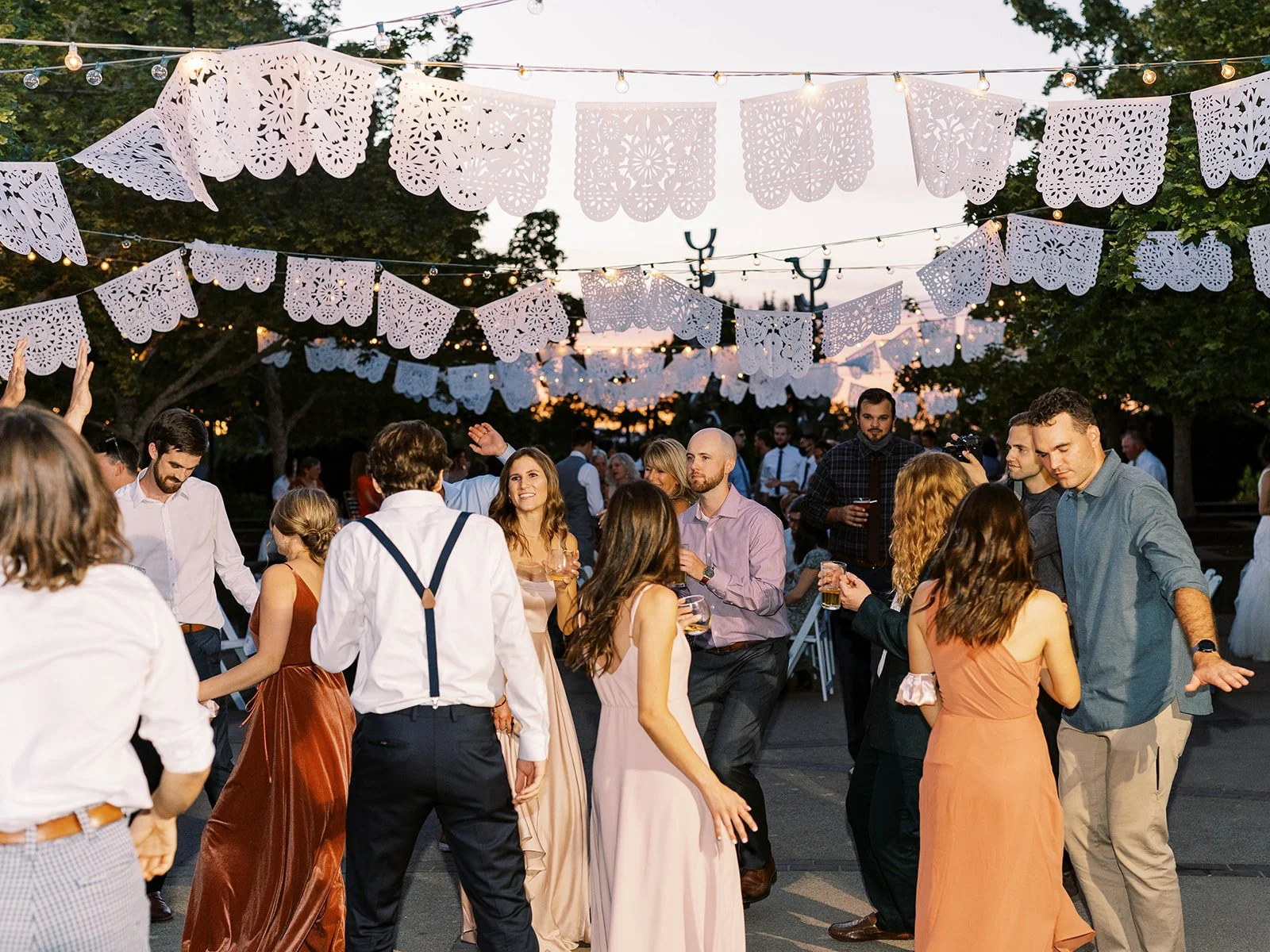 Wedding guests dancing outdoors at sunset beneath string lights and decorative paper banners