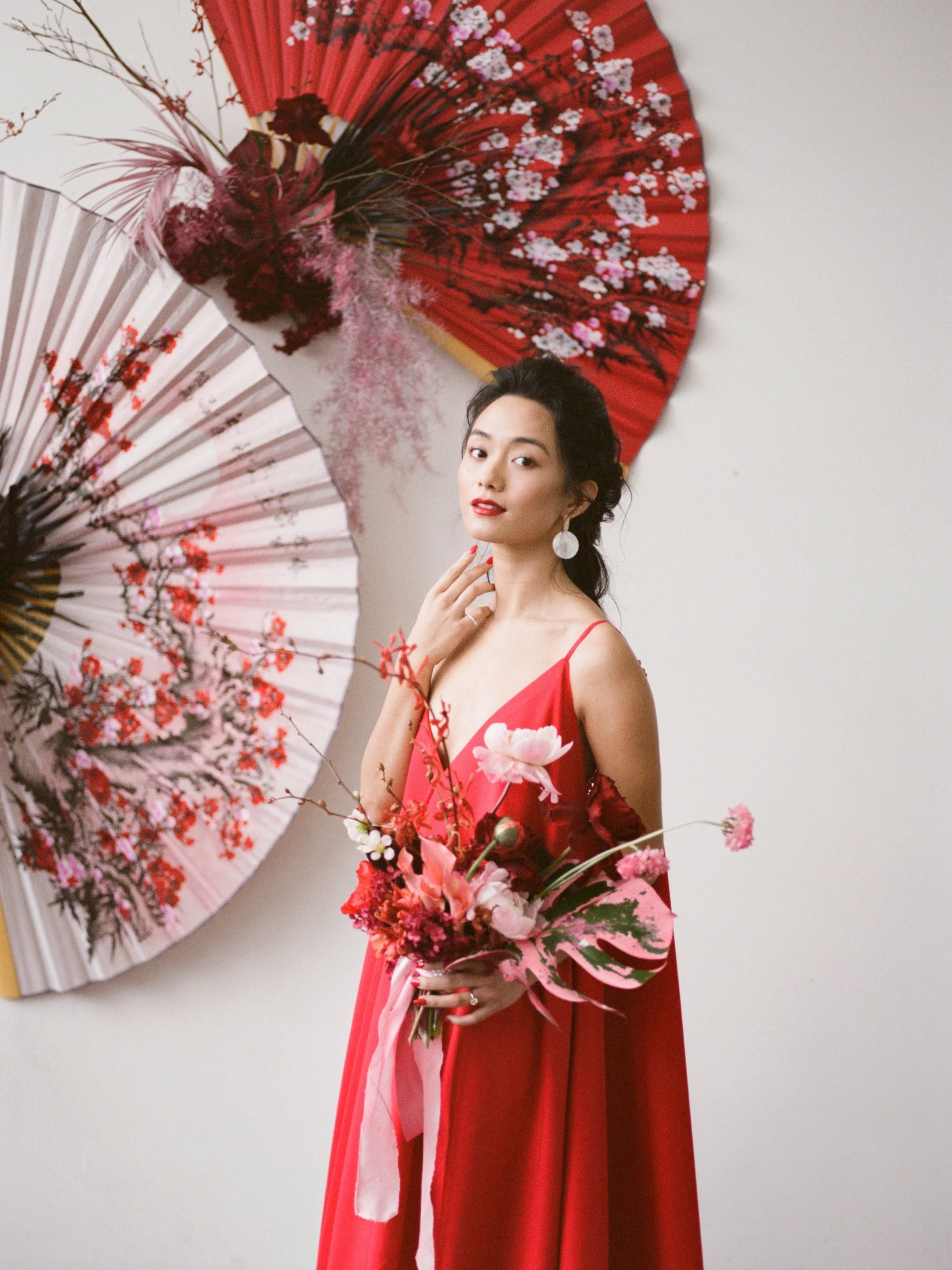 Woman in a flowing red gown holds a bold pink and red bouquet beside decorative Chinese fans.