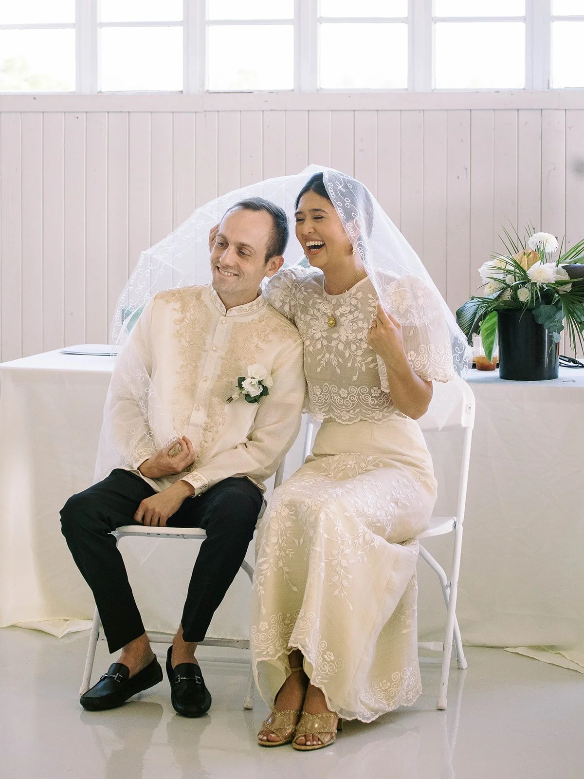 Bride laughing joyfully while seated beside the groom under the traditional Filipino wedding veil during the ceremony
