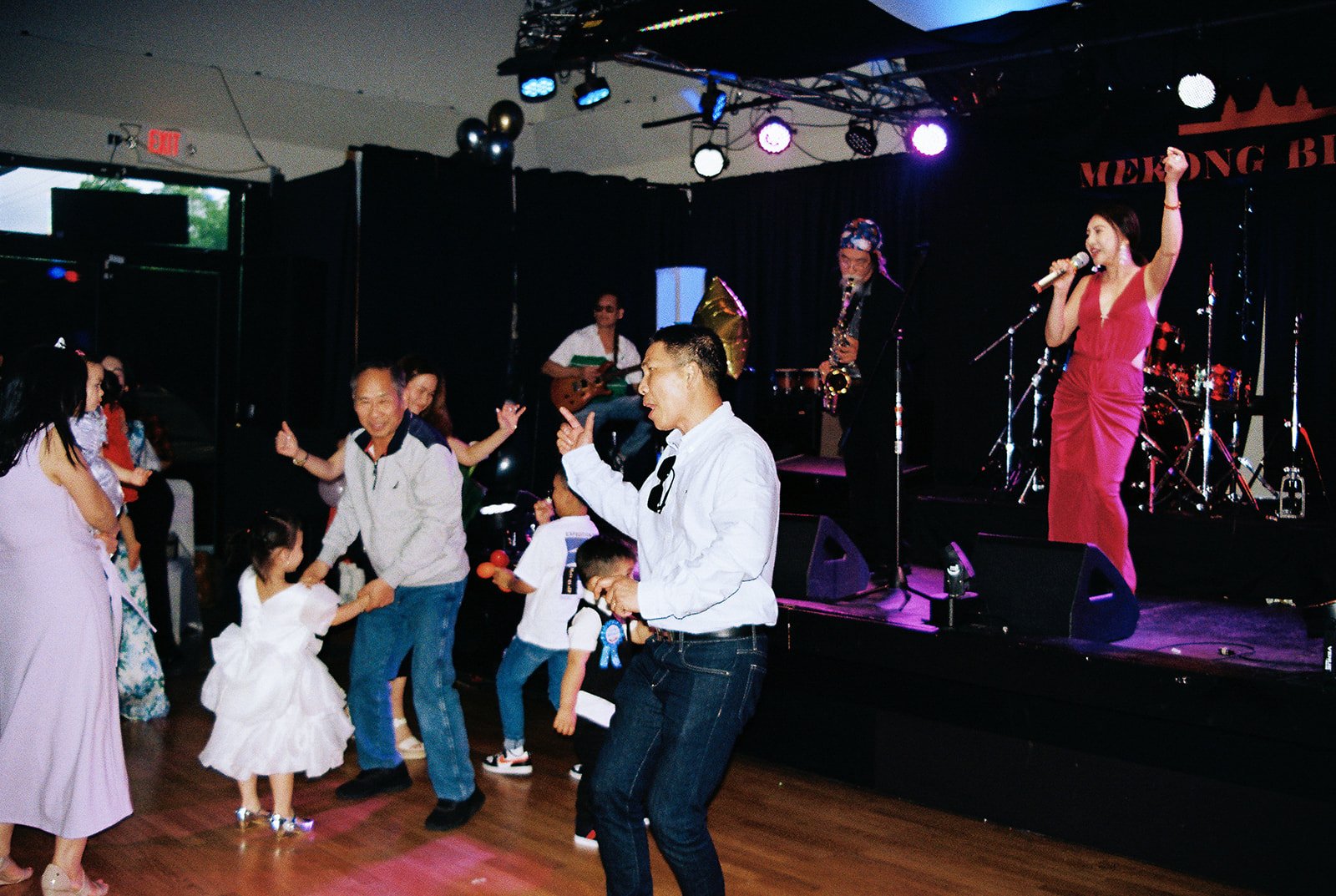 Guests of all ages dancing on the Mekong Bistro dance floor while the live band performs on stage, including a flower girl in a white dress and the saxophonist in the background
