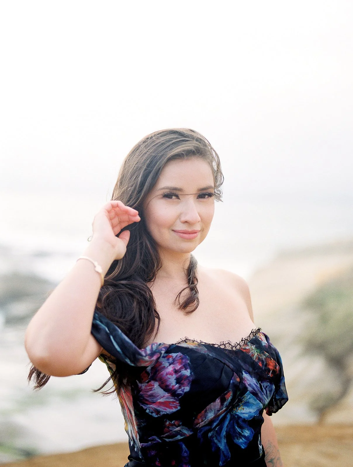 Woman in off-shoulder floral dress standing on coastal cliff, wind blowing hair, soft ocean backdrop