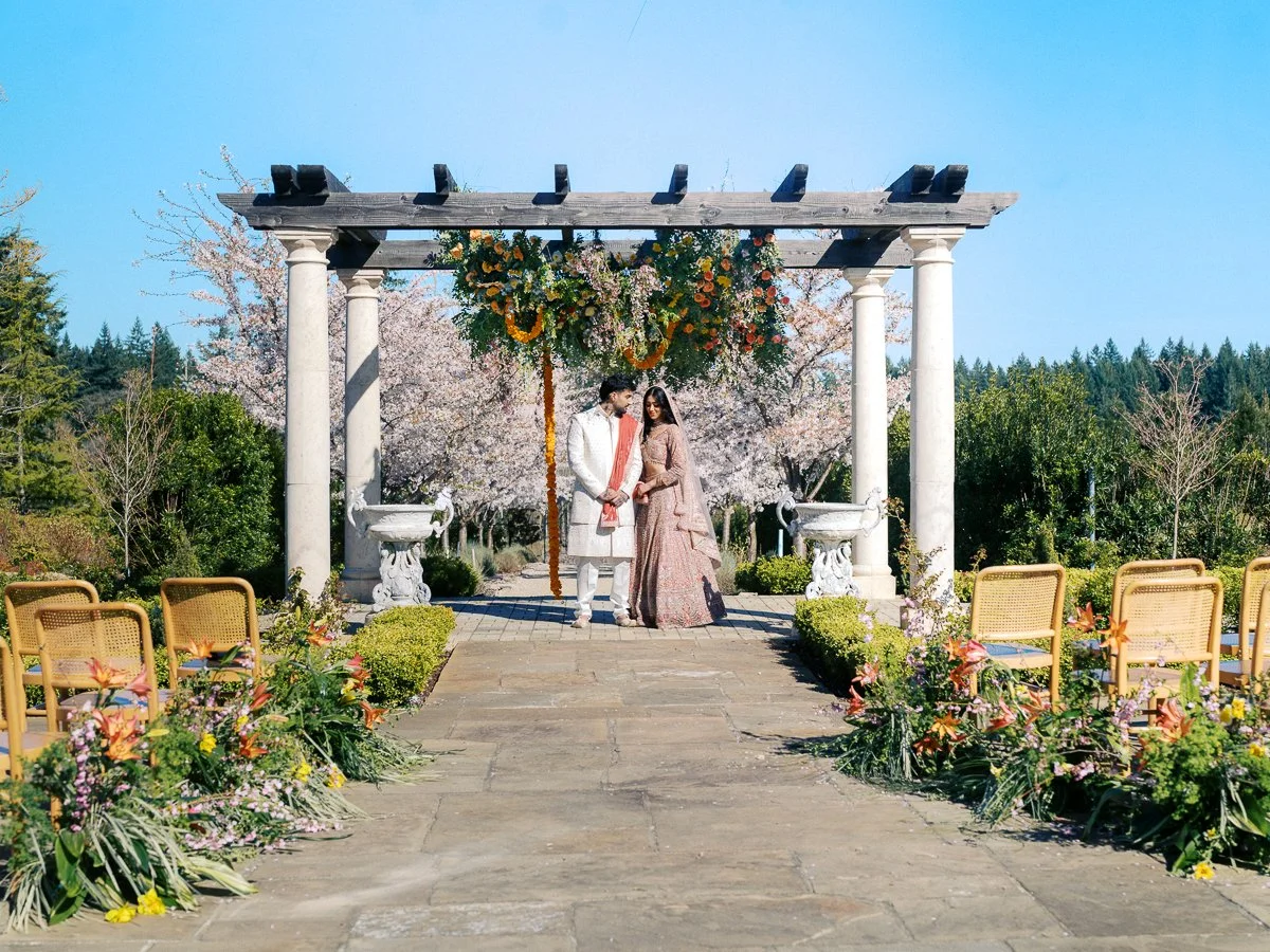 Wide view of couple at an outdoor South Asian wedding ceremony pergola with floral installations and seated guest chairs at Chateau de Lis
