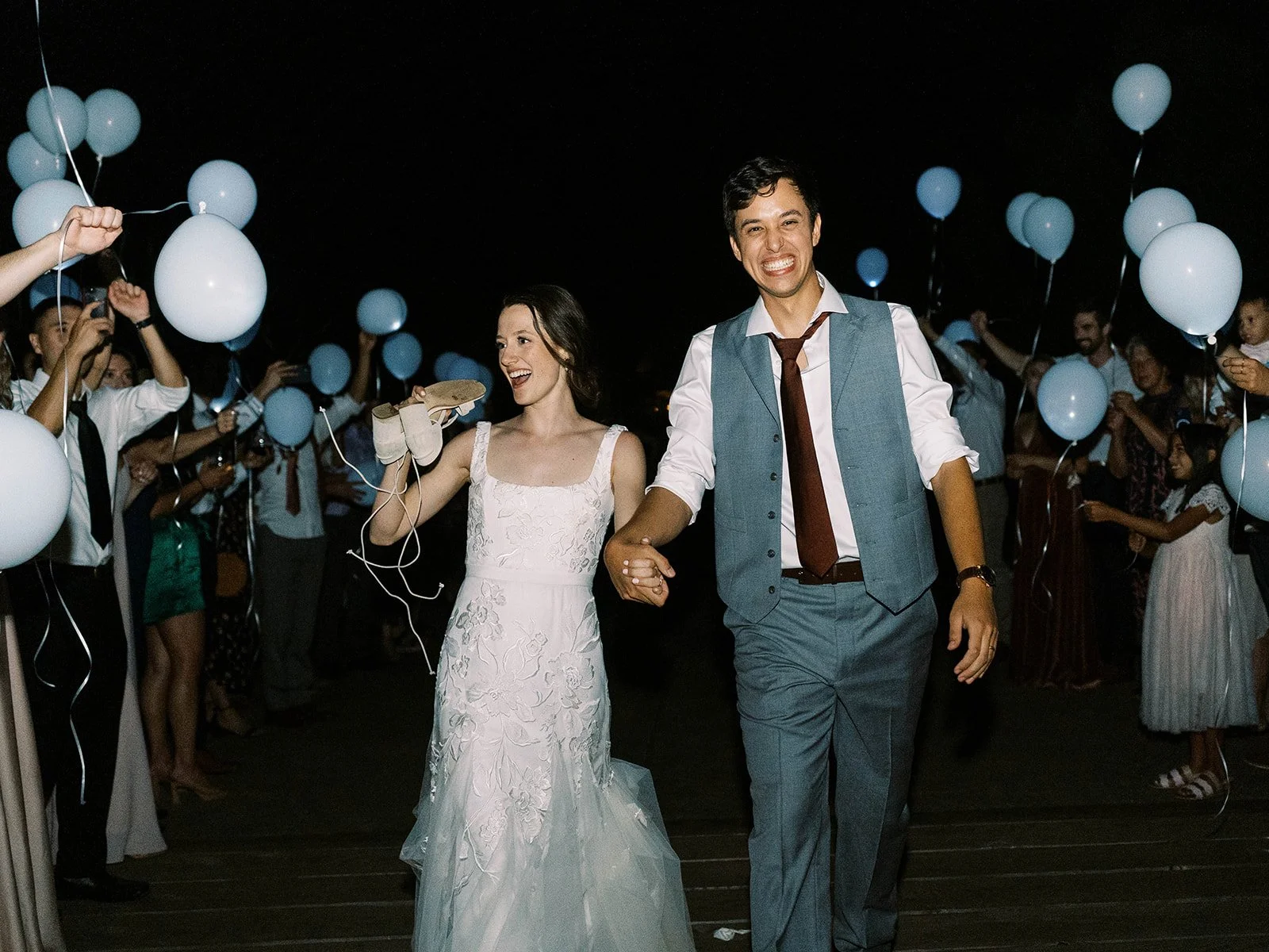 Bride and groom smiling and walking hand in hand with guests holding balloons at night