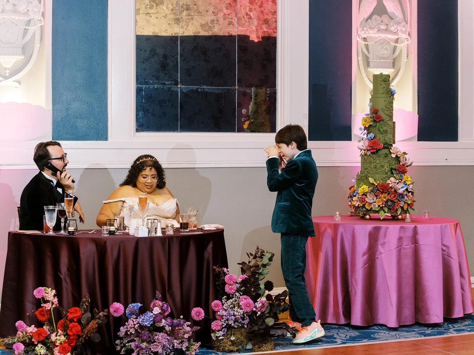 Child in green suit taking a photo of bride and groom seated at reception table with tall floral wedding cake.