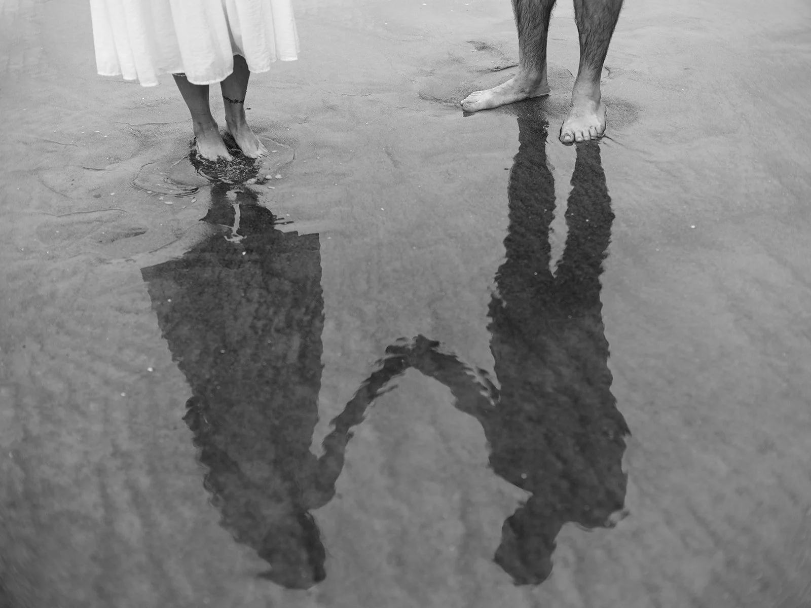 Black and white reflection of a couple holding hands in shallow water on the beach.