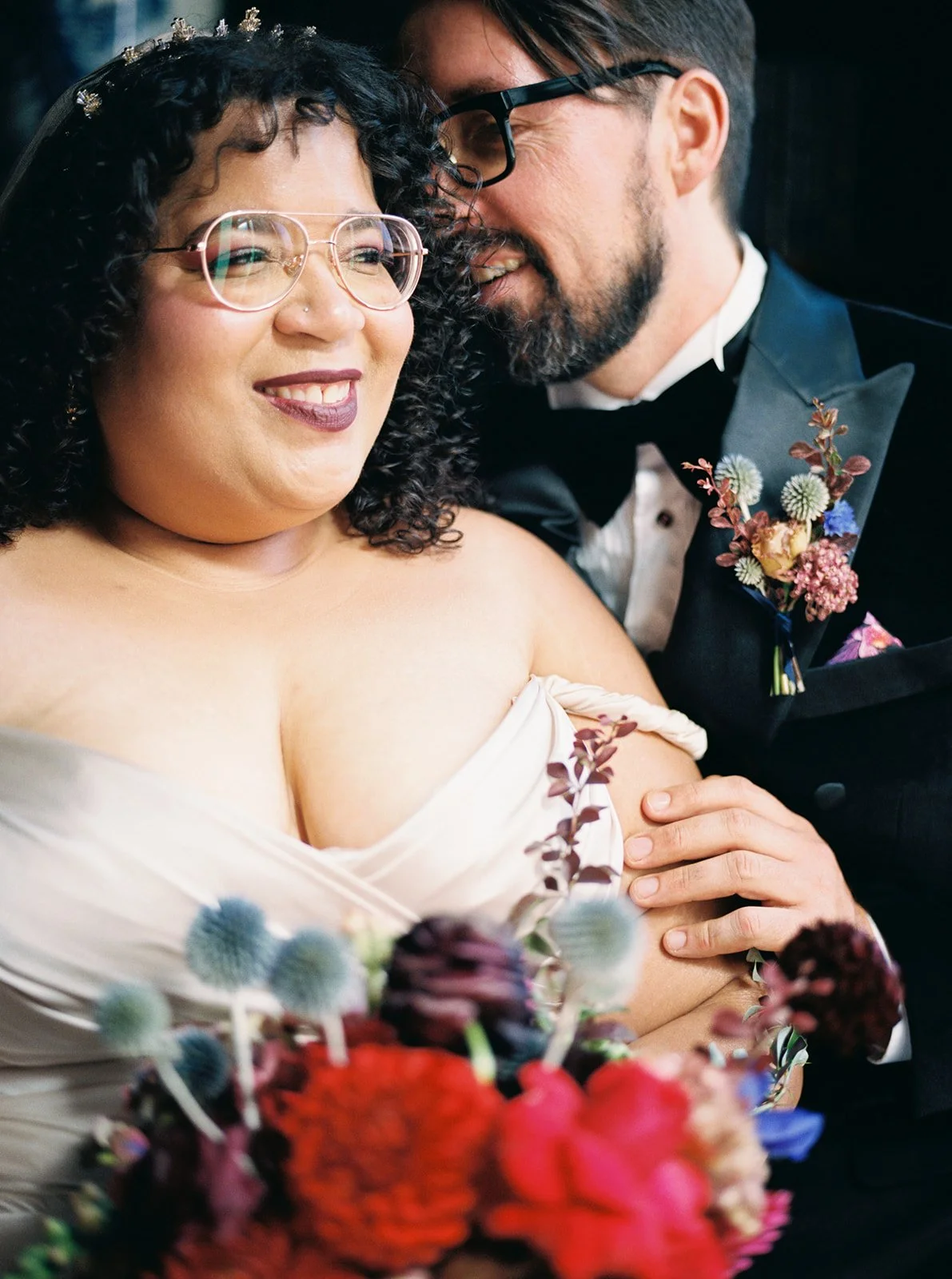 Close-up of couple embracing, groom leaning in as bride smiles, with colorful bouquet in foreground.