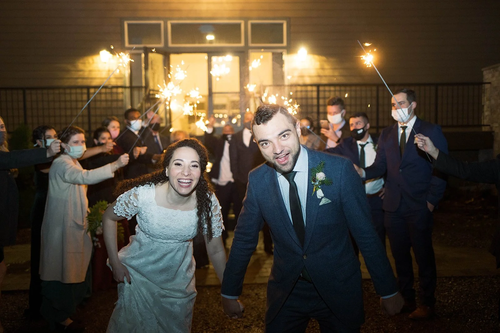 Newlyweds hold hands while running through a nighttime sparkler exit surrounded by wedding guests.
