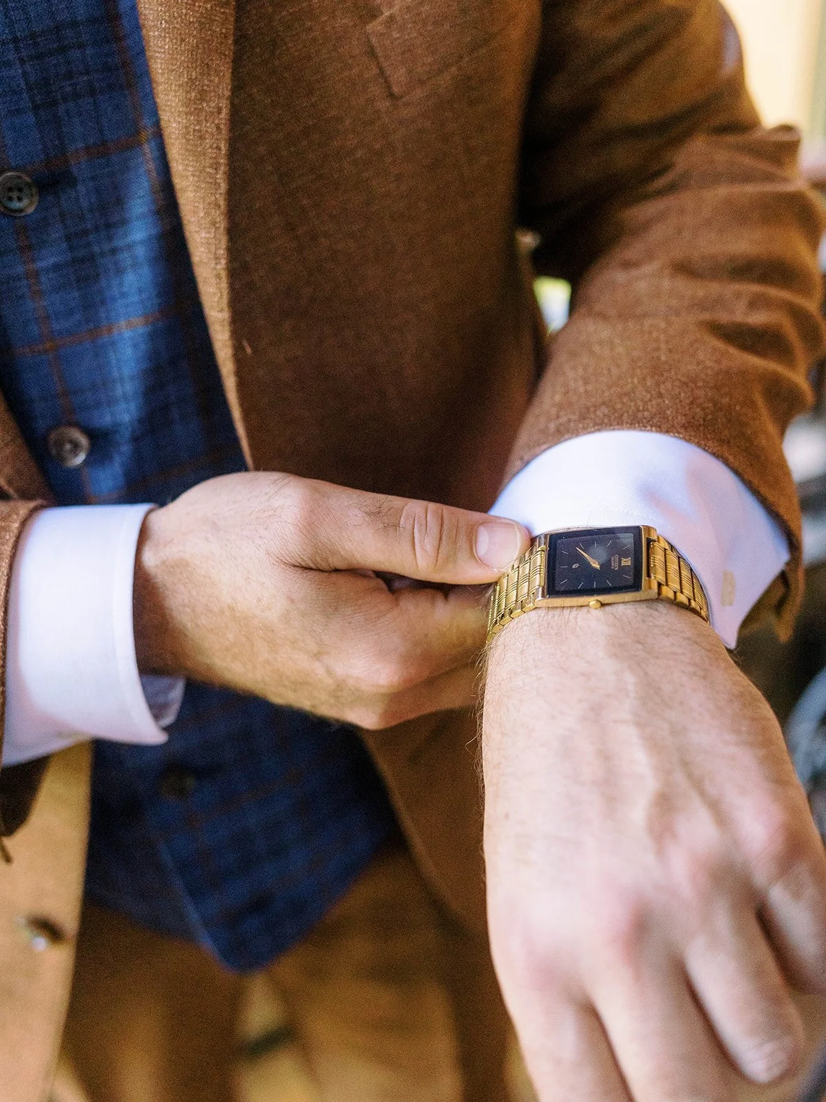 Groom fastening a gold wristwatch while wearing a brown suit and blue plaid vest.