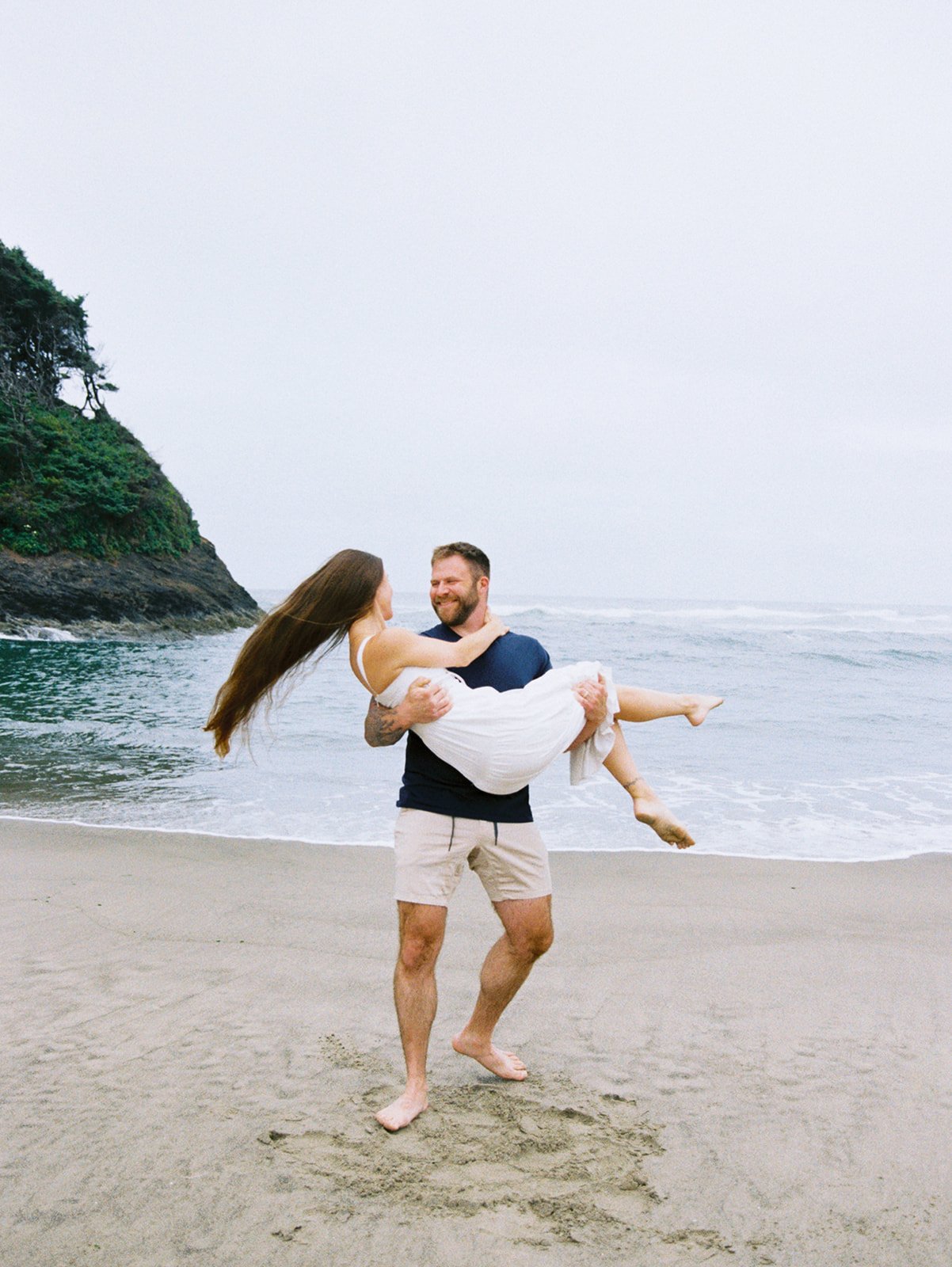 Man lifting woman in white dress on foggy beach with waves and rocky coastline in background.