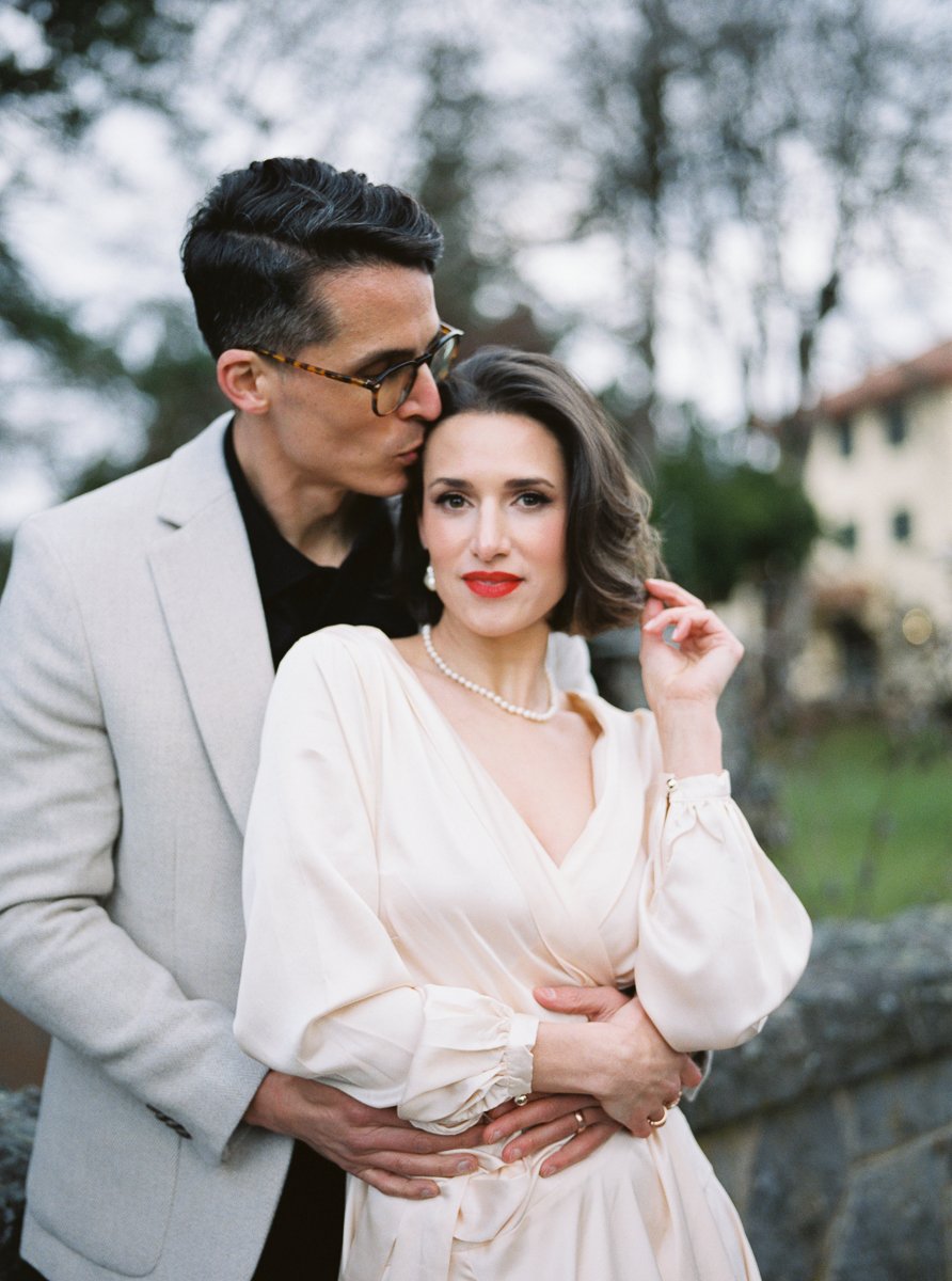 Romantic portrait of couple, man in light blazer kissing woman’s temple, woman in cream dress looking toward camera