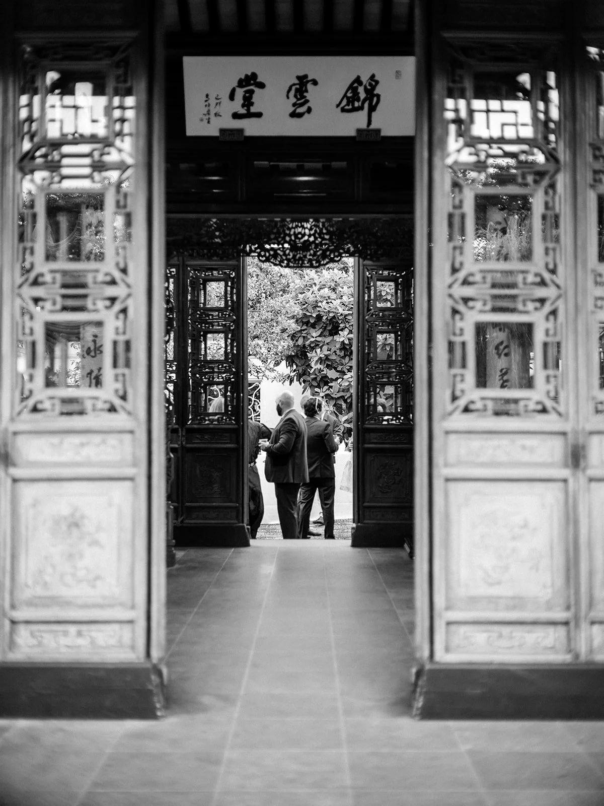 Black and white view through elaborately carved double doors with a calligraphy plaque above, two suited guests silhouetted against the bright garden courtyard beyond.
