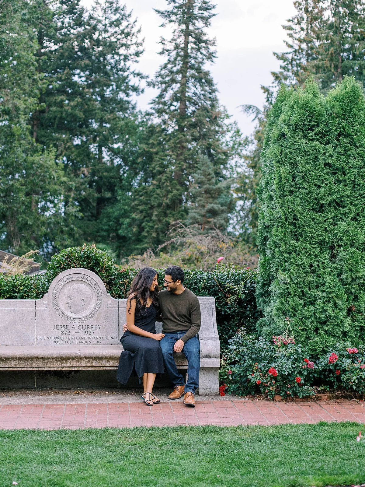 Engaged sit together on the Jesse A. Currey memorial bench surrounded by roses and evergreen trees.