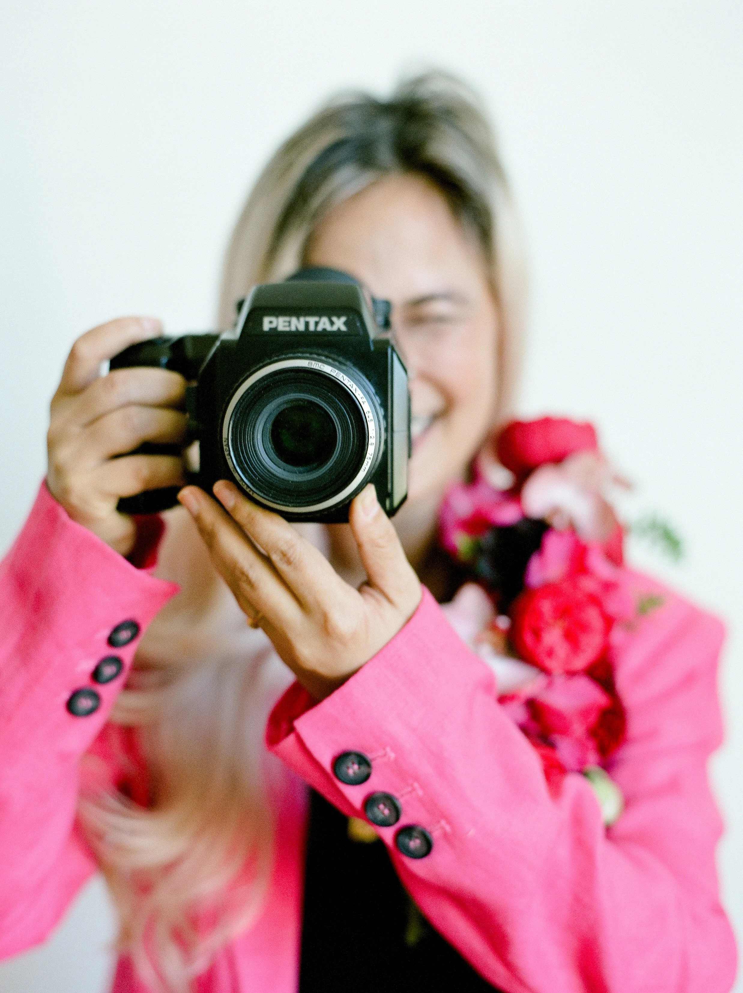 Mylyn Wood holding a Pentax film camera while wearing a bright pink blazer and colorful floral lei.