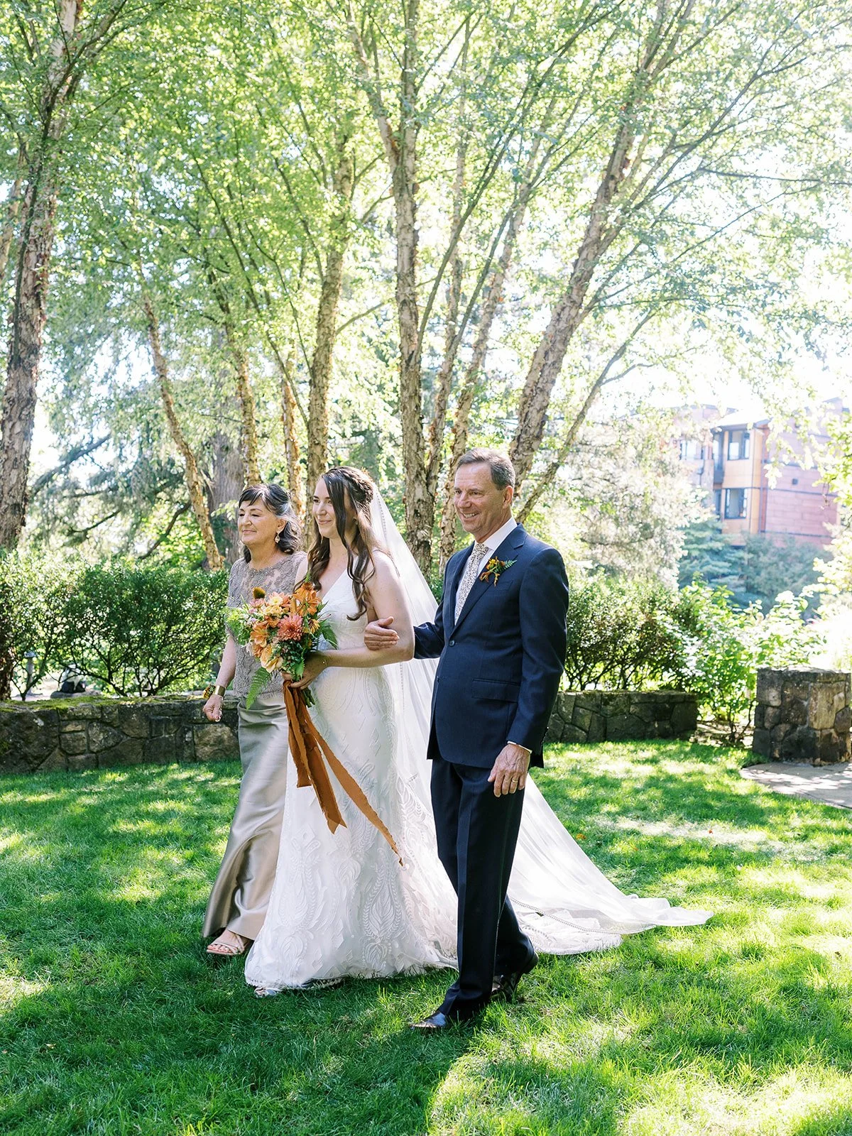Bride walking down the aisle with two adults on a grassy lawn, holding a bouquet with warm-toned flowers.