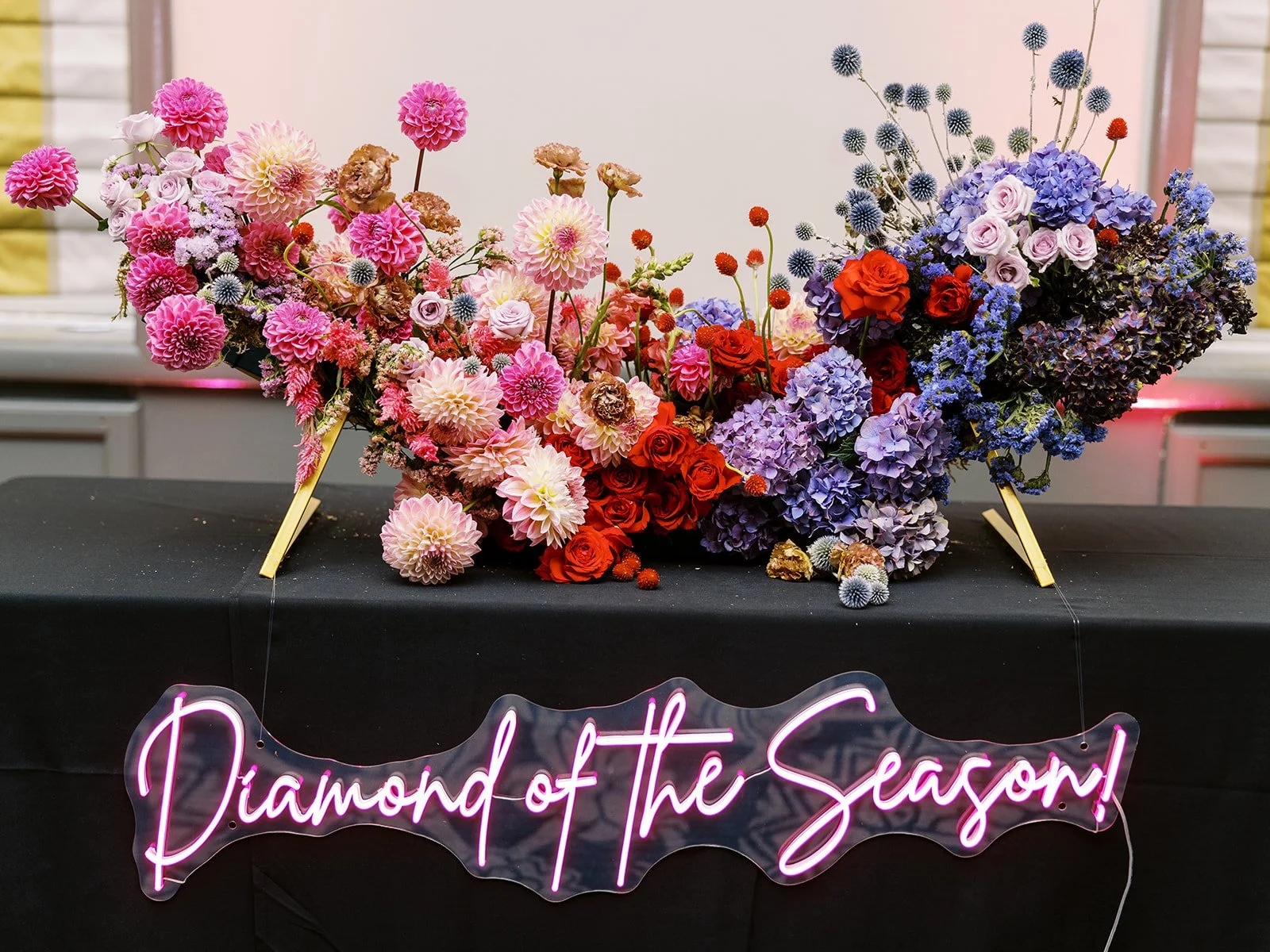 Wide floral installation in pink, red, and purple tones above neon “Diamond of the Season” sign.
