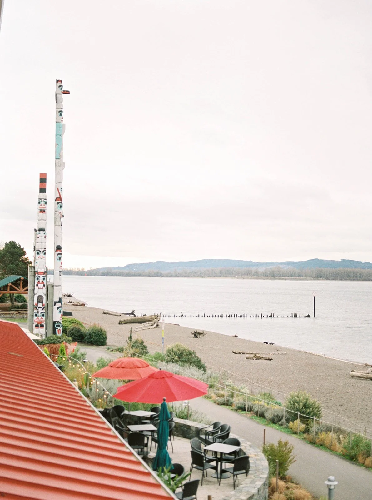Riverfront Oregon wedding setting with totem poles, red umbrellas, beach, and distant wooded hills.
