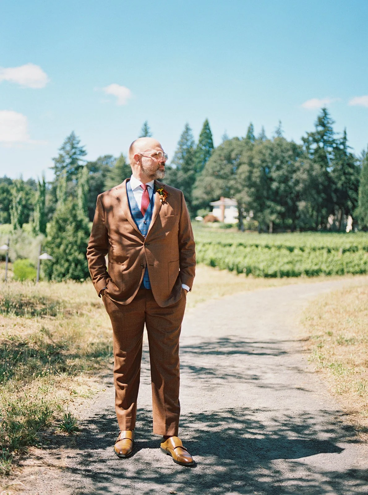 Groom in brown suit looking to the side on outdoor path with vineyard scenery