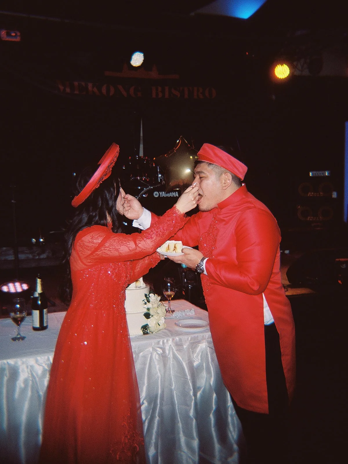 Bride and groom in matching red Vietnamese áo dài feeding each other wedding cake at Mekong Bistro, a Yamaha drum kit visible on stage behind them