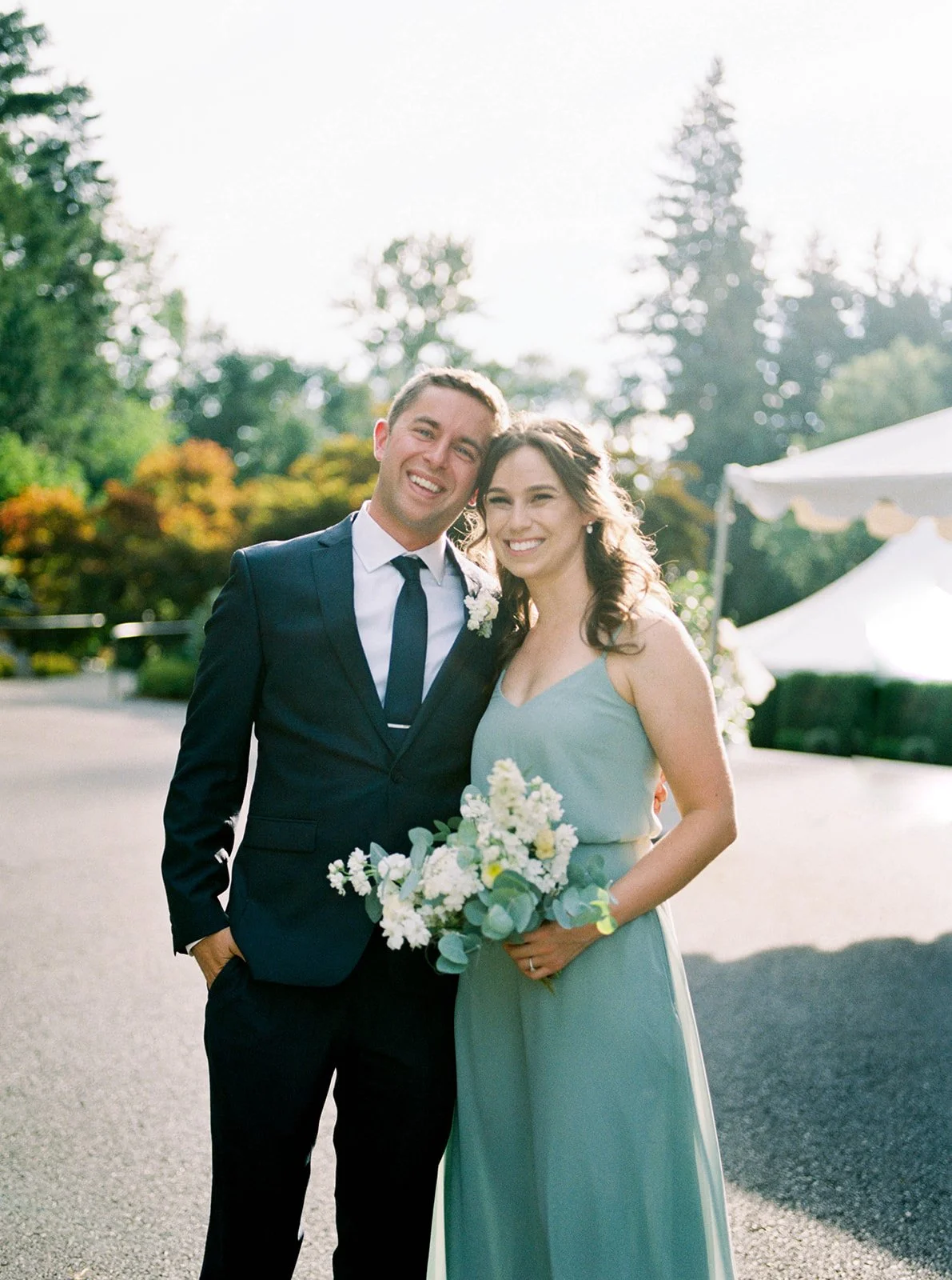 Bridesmaid and groomsman pose outdoors in formal wedding attire, holding a bouquet in soft evening light.