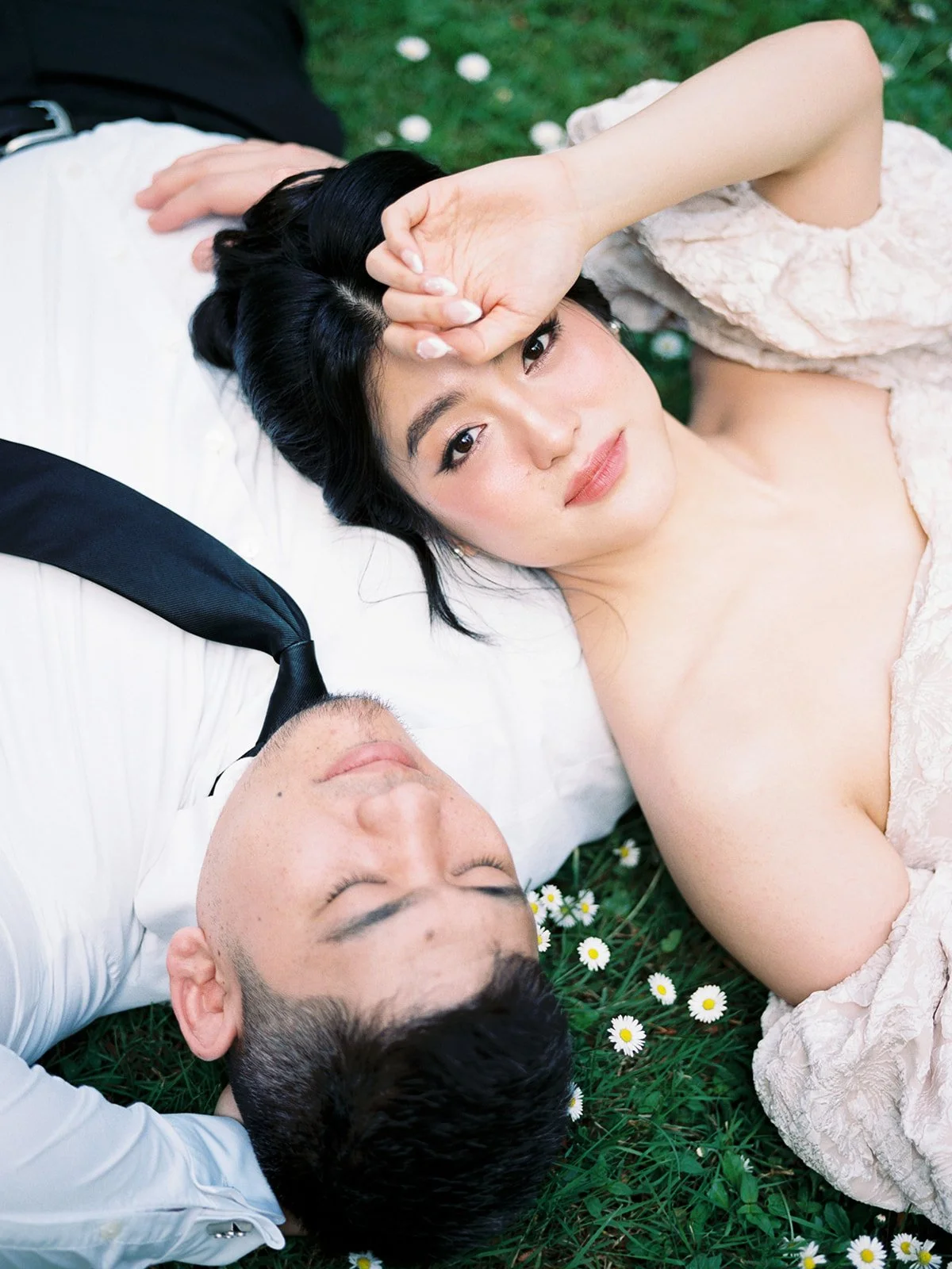 Overhead close-up of the bride and groom lying on a daisy-covered lawn at The Grotto, she gazing up at the camera, he with eyes closed