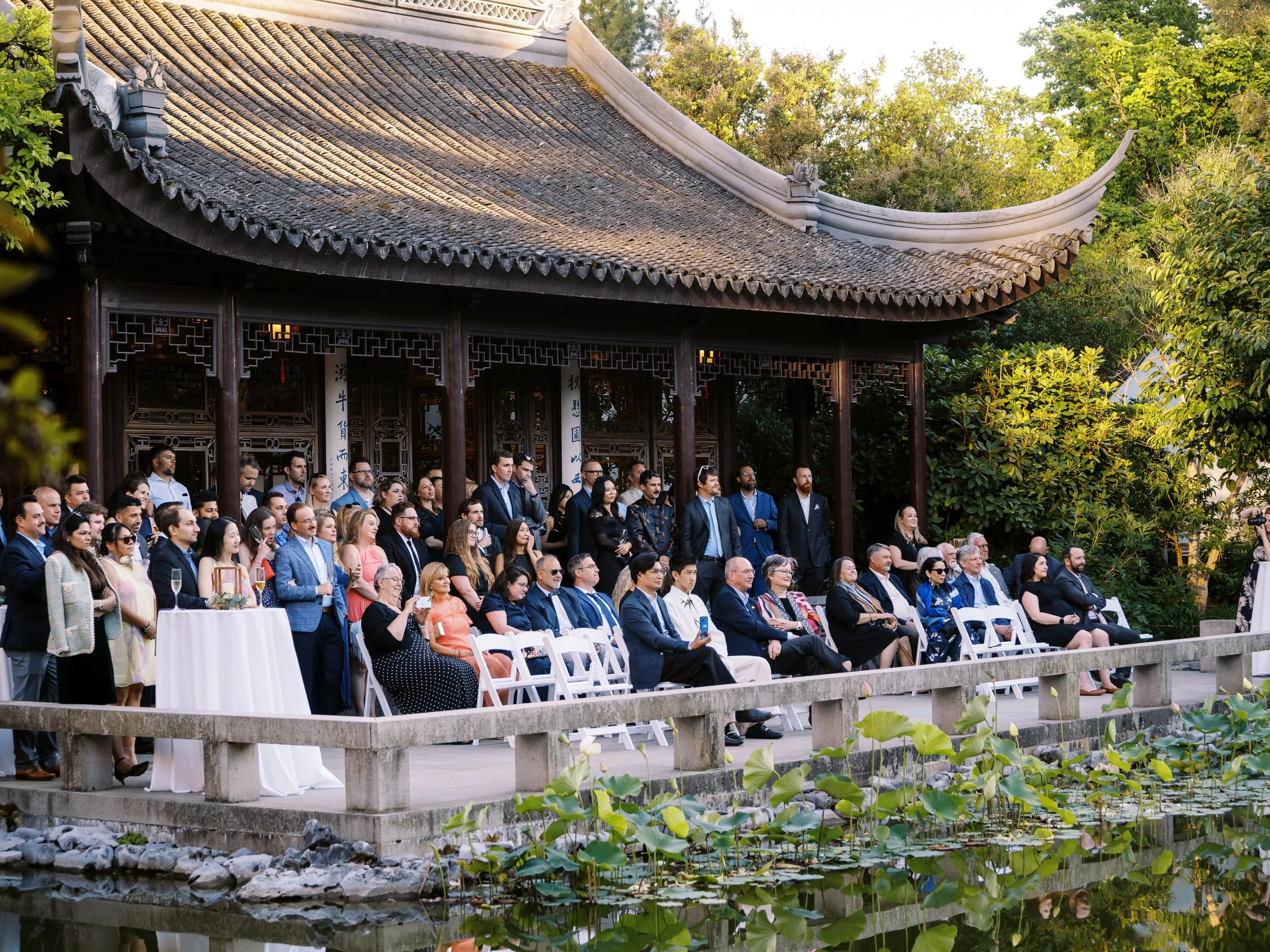 A seated audience watches a wedding ceremony outside a traditional Chinese pavilion beside a lotus-filled pond.