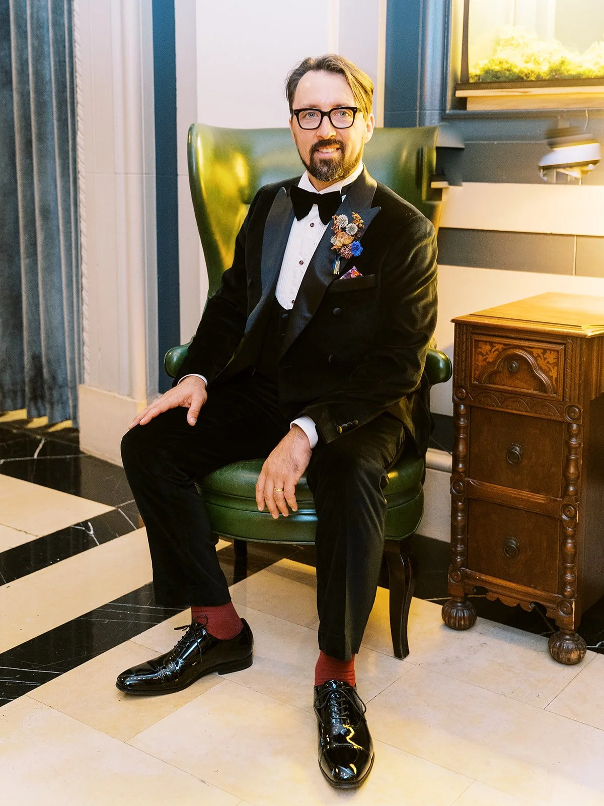 Groom seated in green leather chair wearing black tuxedo, posed indoors beside wooden side table.
