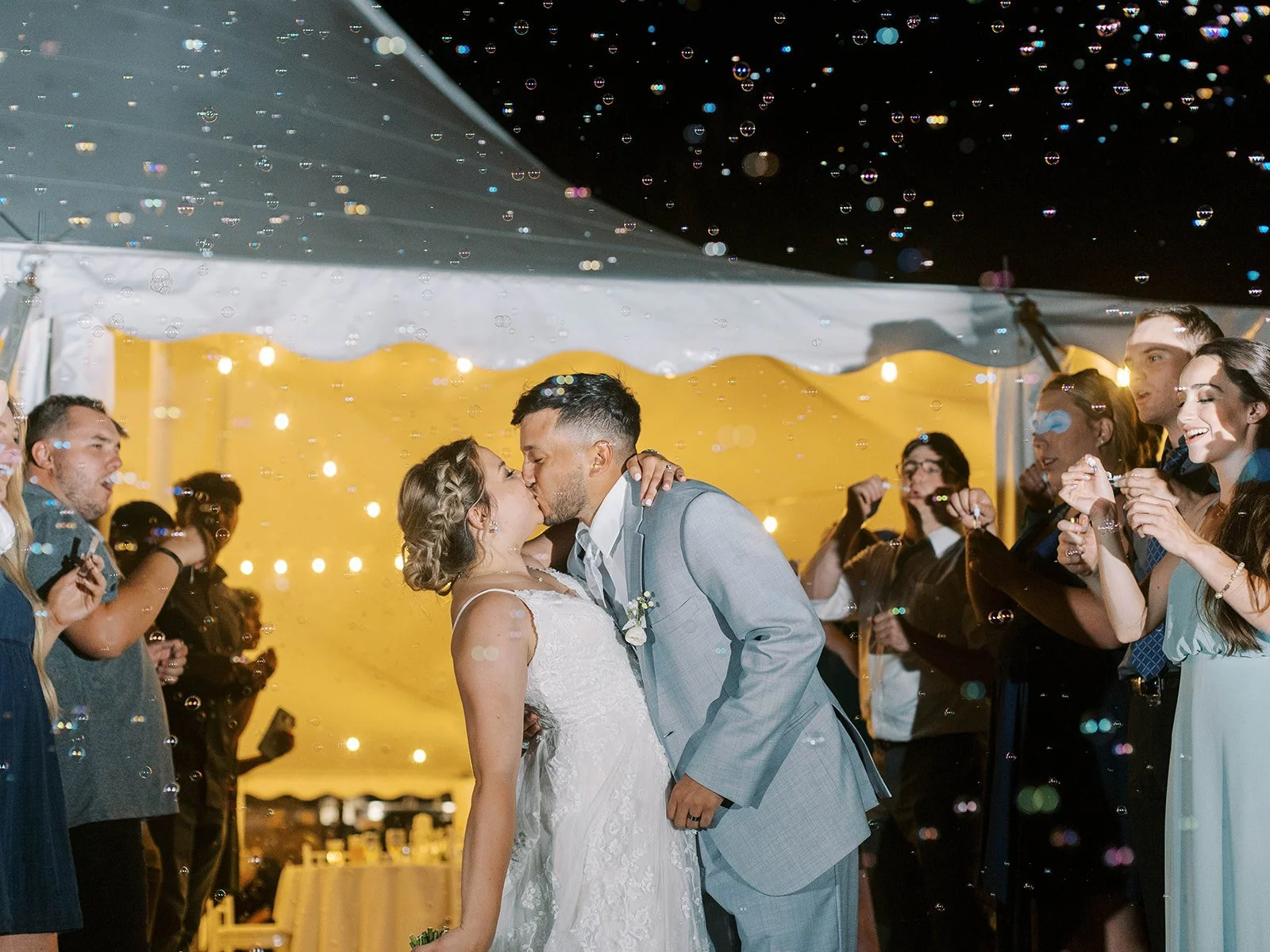 Bride and groom share a kiss during their nighttime bubble send-off, framed by wedding guests and glowing tent lights.