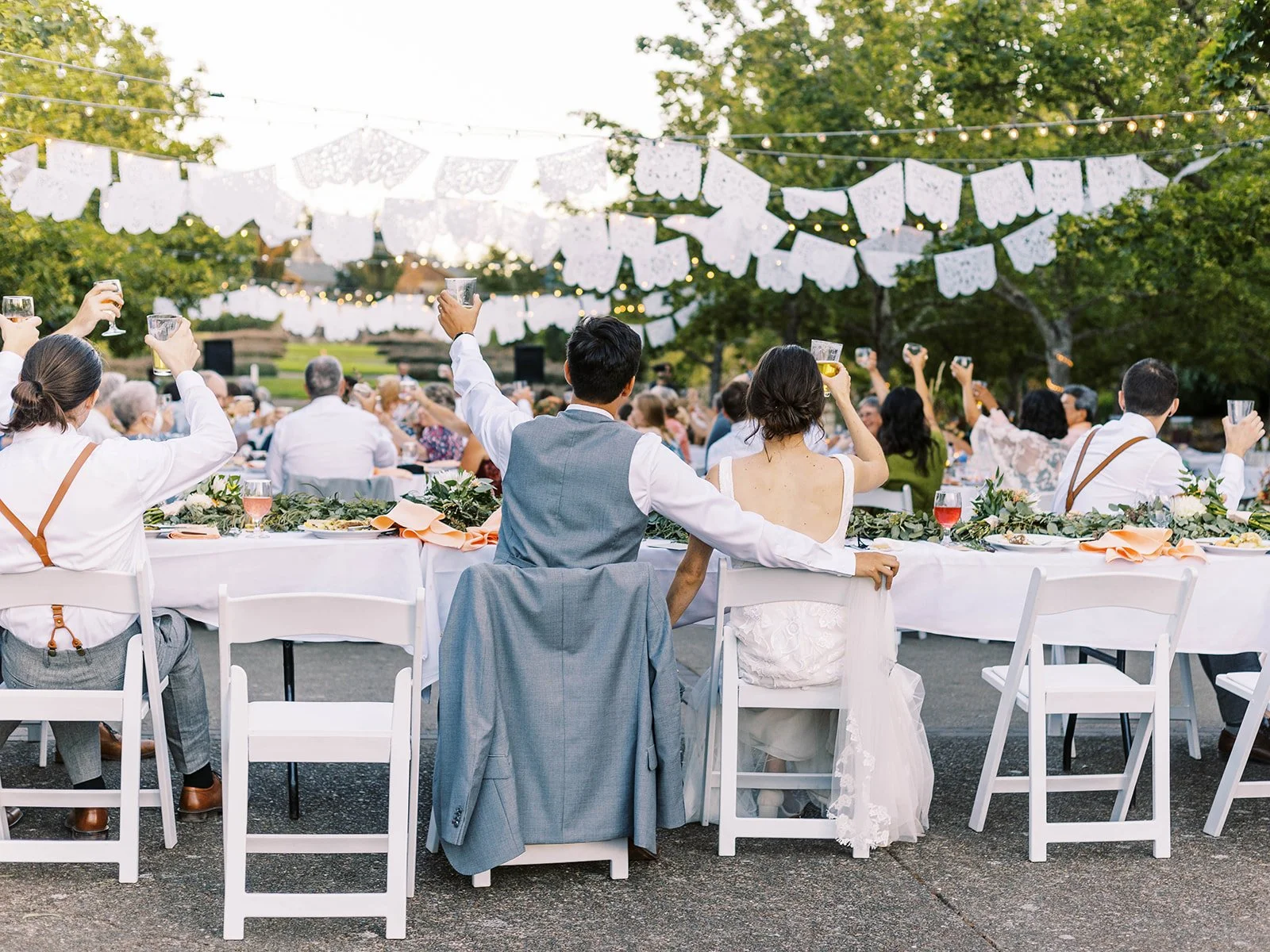Wedding guests raising glasses in a toast at outdoor reception with string lights and decorative banners