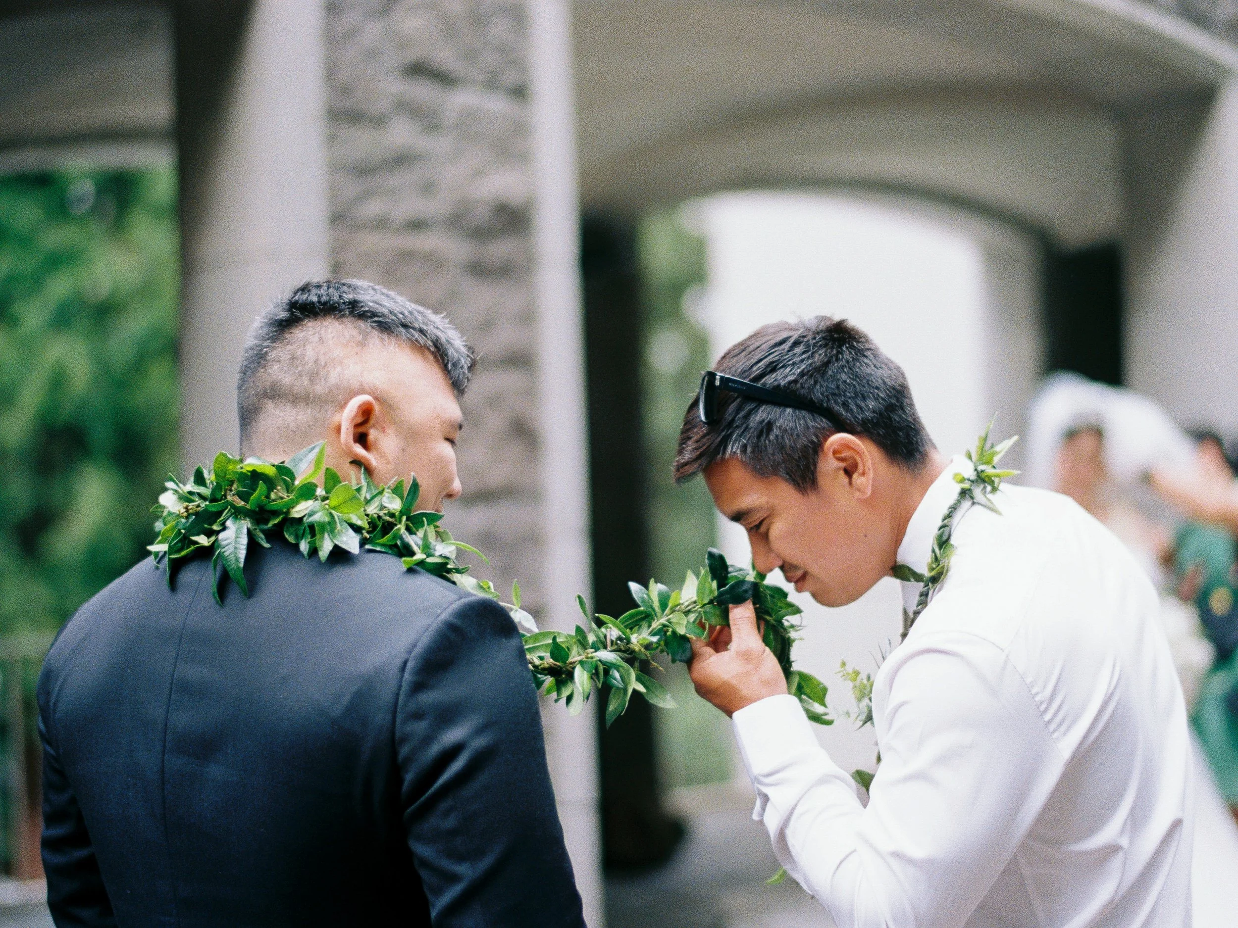 Wedding guest lifts a green lei to his face while standing beside another man wearing a matching lei.