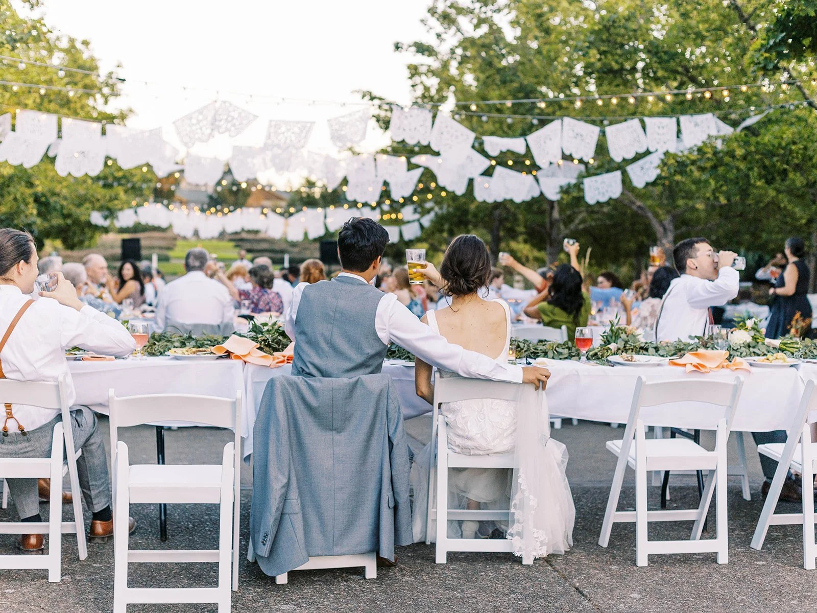 Newlyweds seated together at an outdoor reception beneath papel picado and string lights at The Oregon Garden.