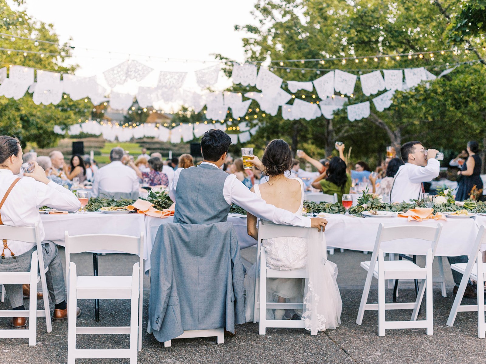 Bride and groom seated with guests at a long outdoor wedding reception table beneath string lights and white papel picado banners.