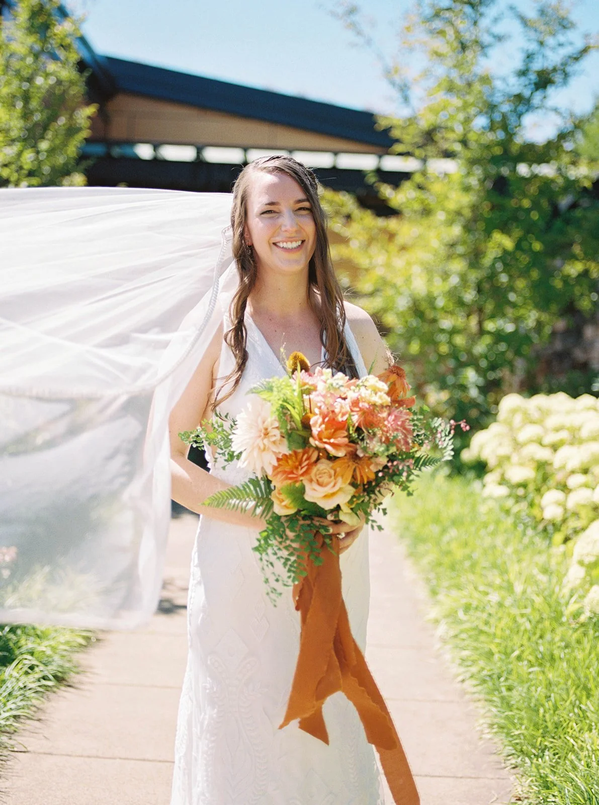Smiling bride holding vibrant bouquet with long ribbon, veil flowing in sunlight