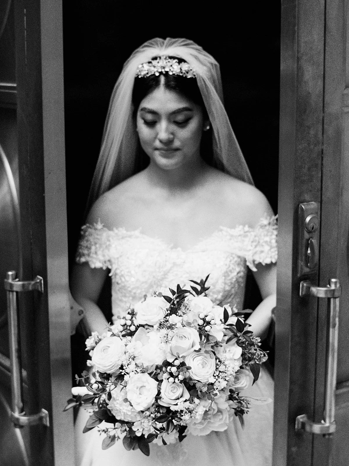 Black and white portrait of the bride framed between the gold doors of The Grotto's chapel, looking down at her white rose and greenery bouquet
