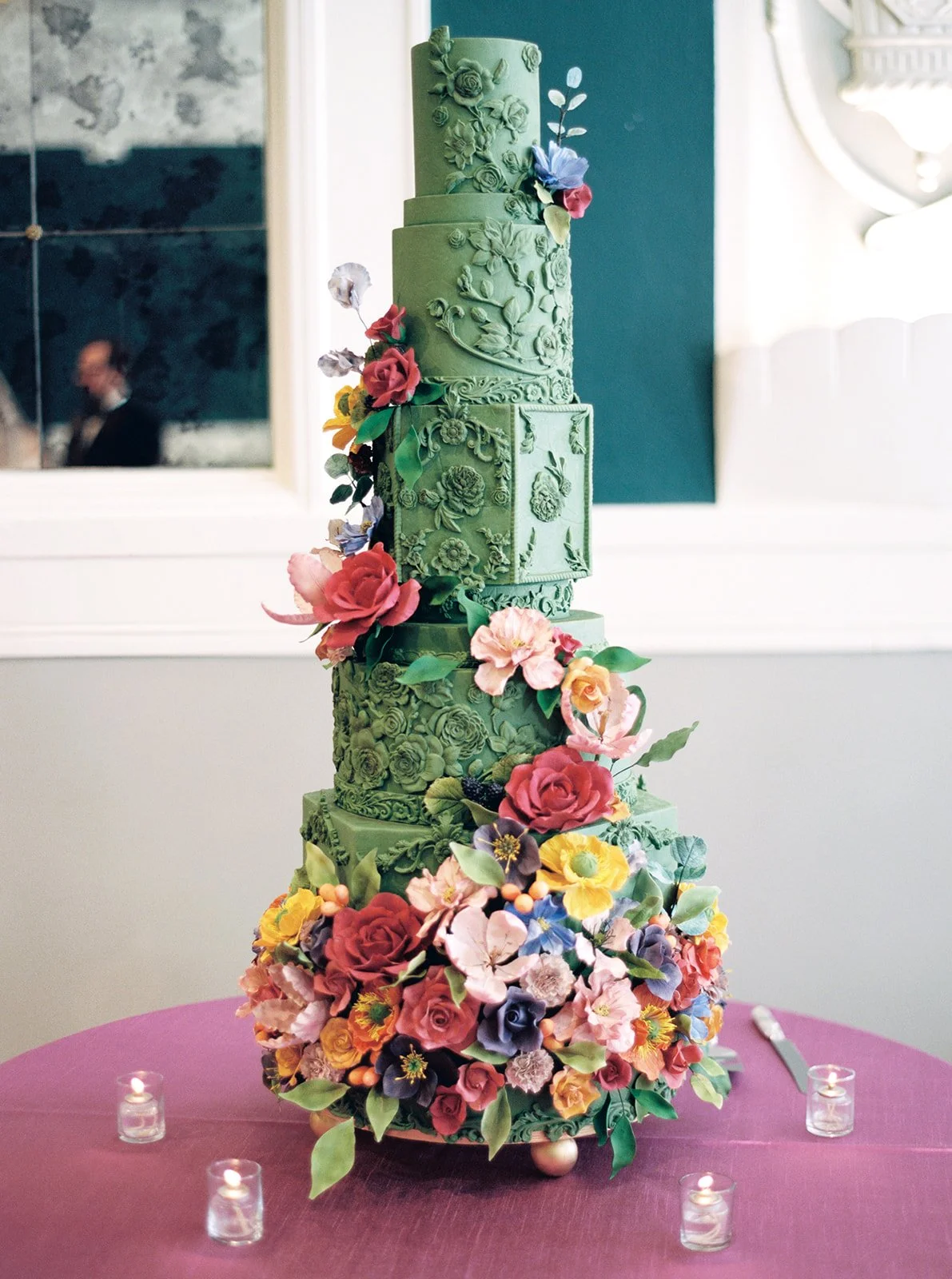 Tall green wedding cake with ornate detailing and cascading sugar flowers displayed on a pink linen table with candles.