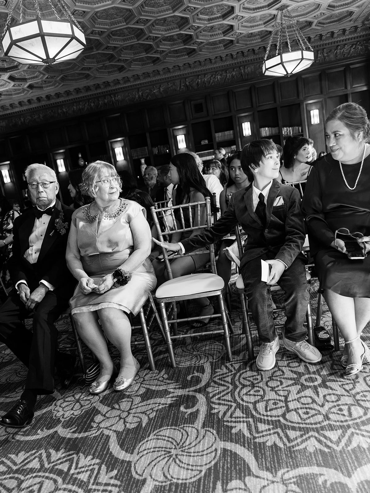 Black and white candid of wedding guests seated in rows inside ornate ceremony space.