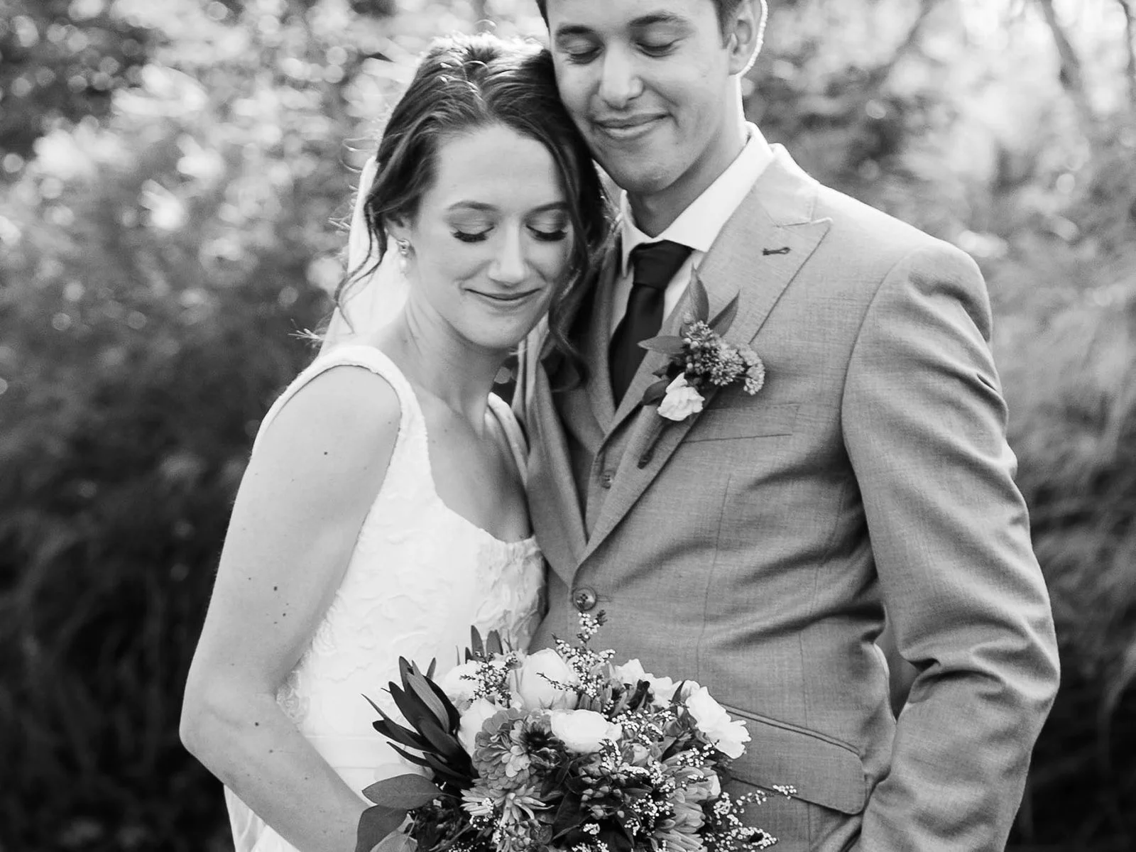 Bride and groom embrace outdoors, holding bouquet, soft natural light, black and white wedding portrait