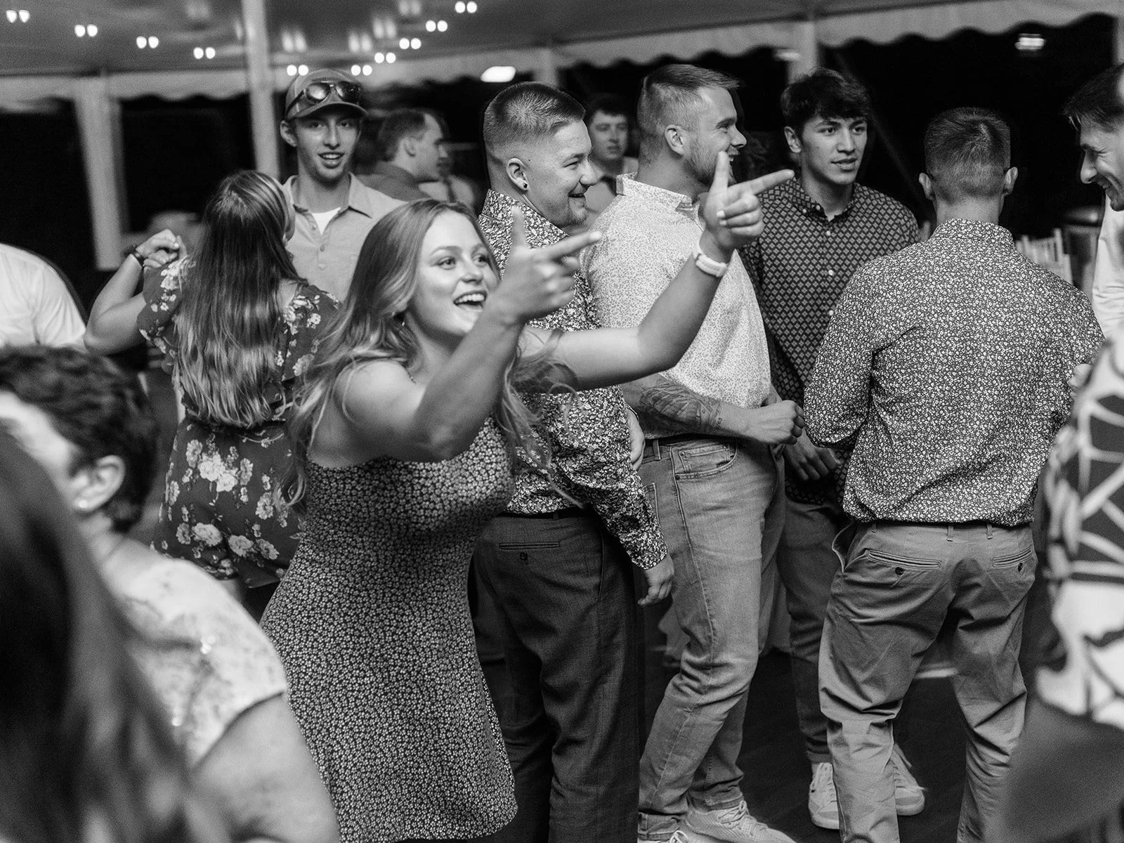 Wedding guests dancing energetically at the reception, with a woman pointing playfully on the dance floor in black-and-white.