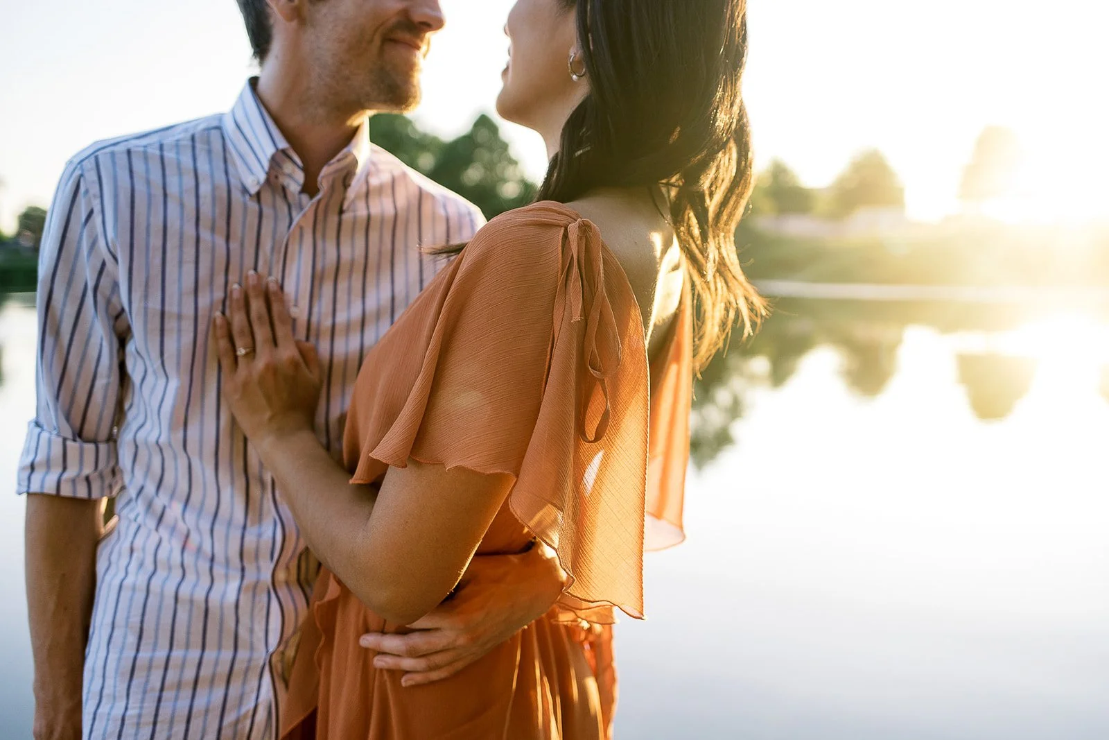 Couple standing close by a lake at sunset, woman in orange dress resting hand on partner’s chest, soft golden light