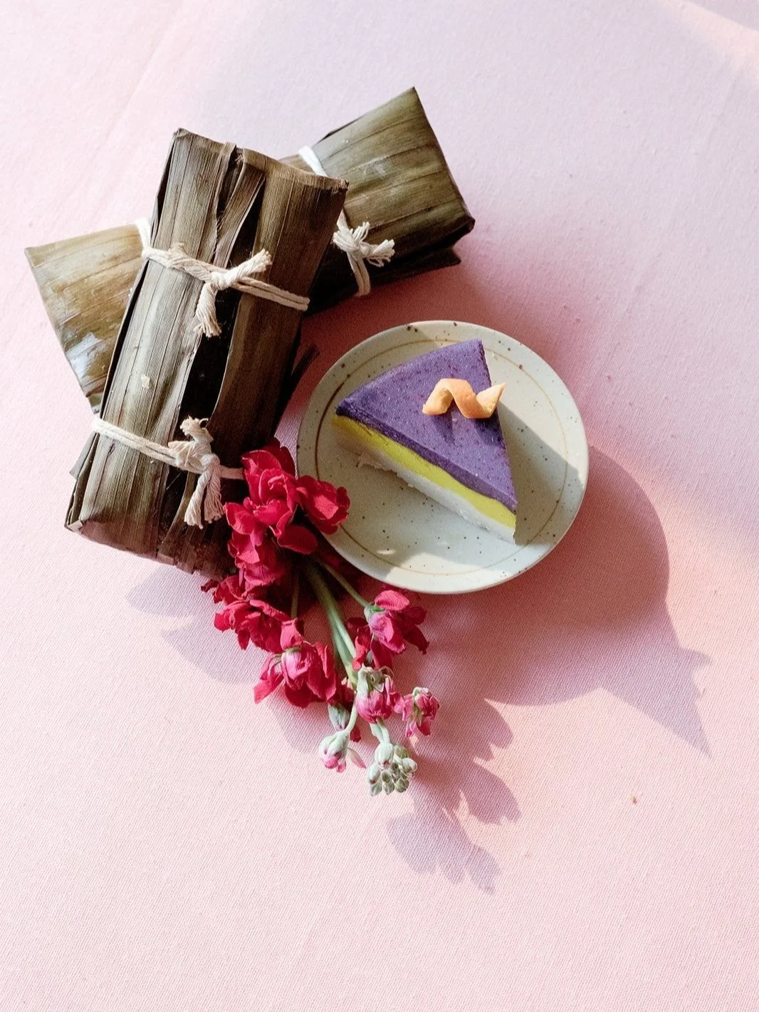 Purple and yellow layered dessert beside wrapped banana-leaf parcels and red flowers