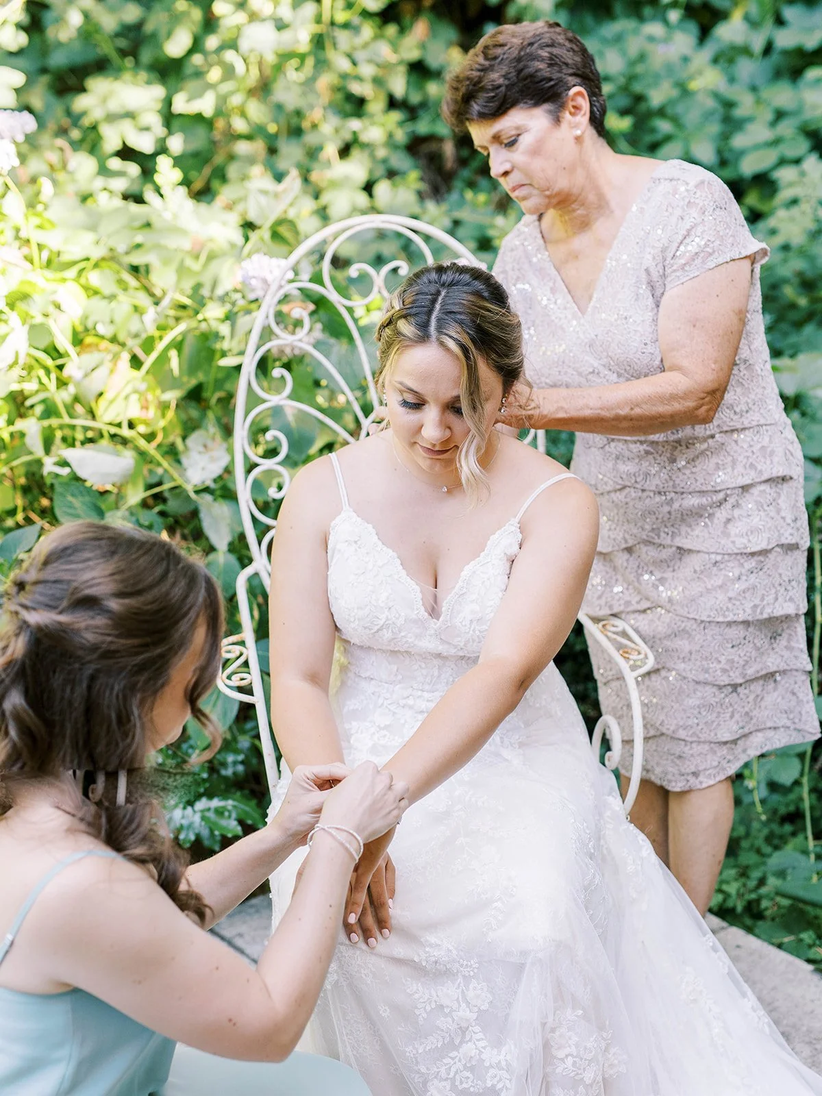 Bride seated in lace gown while mother fastens necklace and bridesmaid adjusts bracelet in garden setting.