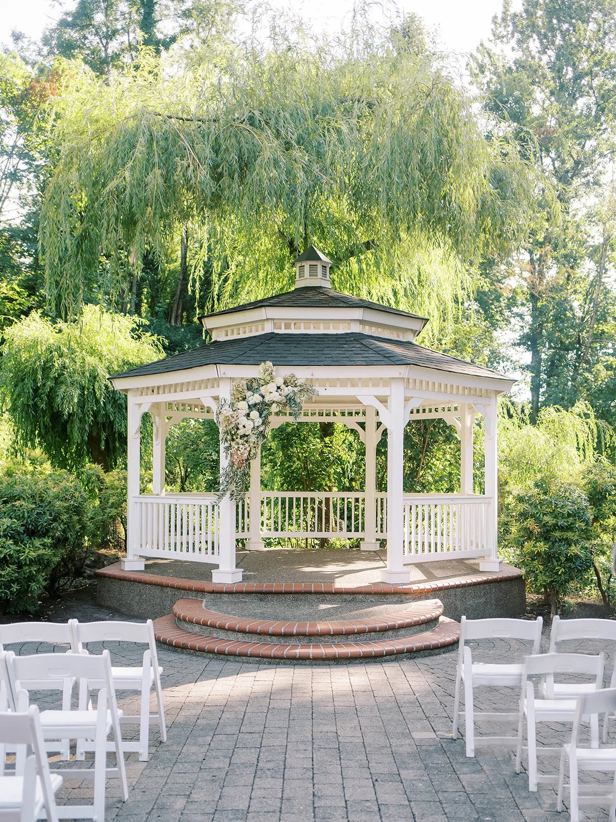 White garden gazebo ceremony site at Abernathy Center’s Abigail’s Garden surrounded by willow trees and lush greenery.