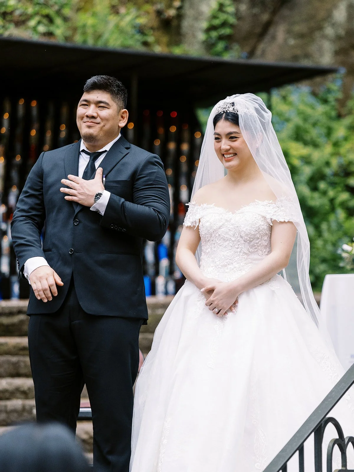 Groom with hand on his chest and a moved expression standing beside the smiling bride in her tiara and veil at The Grotto ceremony site, the lit grotto glowing behind them