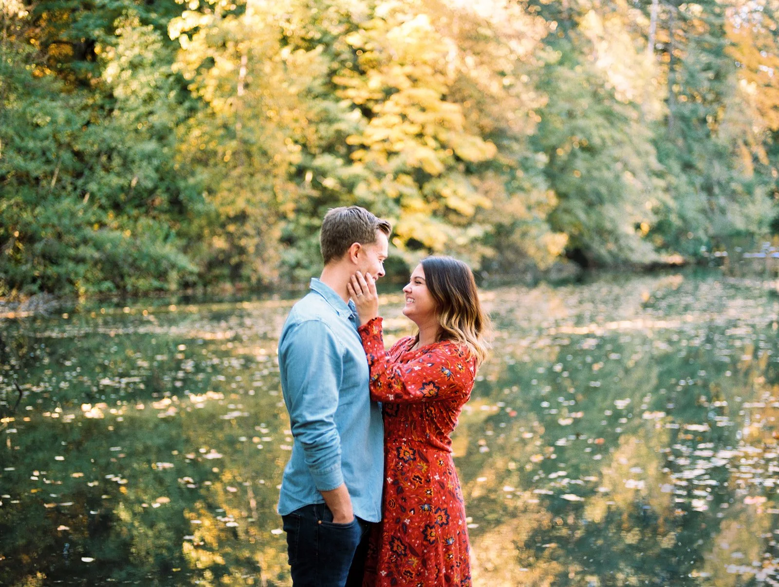 Woman in a red floral dress touching man’s face by a river with golden foliage