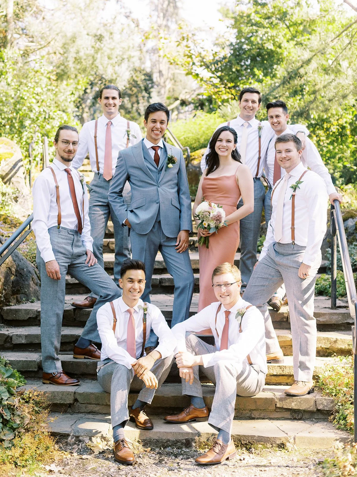 Wedding party posing on garden steps with attendants in gray suits, suspenders and warm neutral dresses.