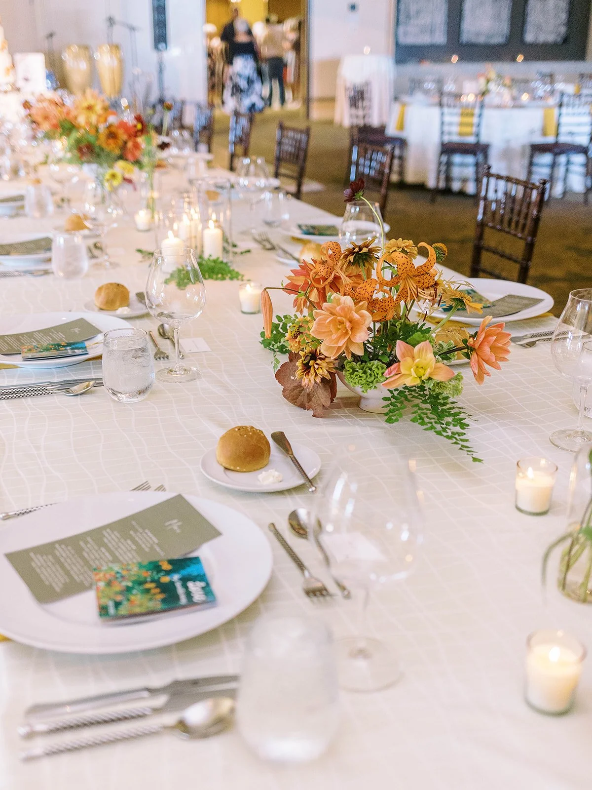 Wedding reception table set with candles, place settings and low arrangements of peach and orange flowers.