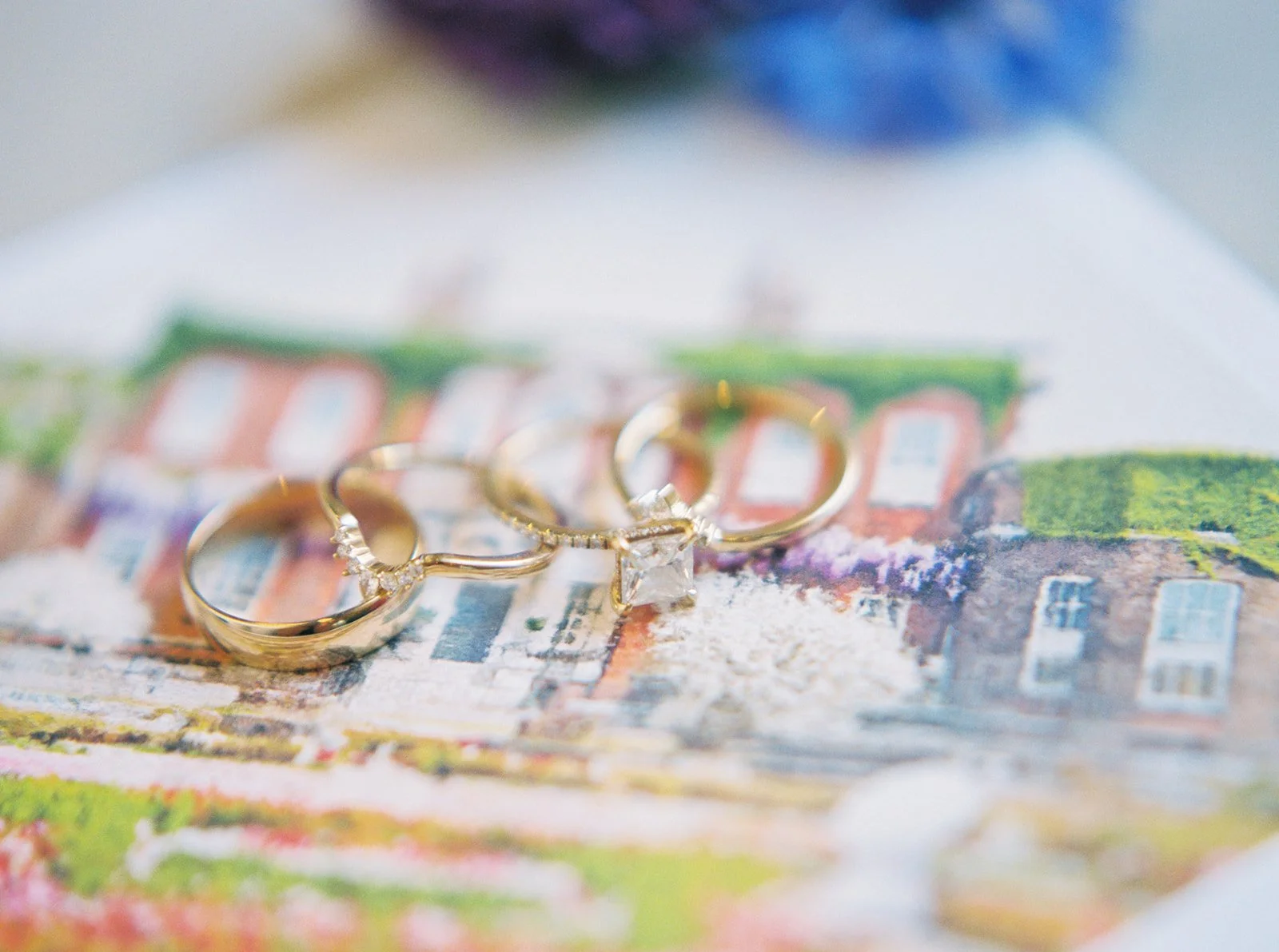 Gold wedding bands and diamond engagement ring resting on an illustrated Sentinel Hotel card, softly blurred.
