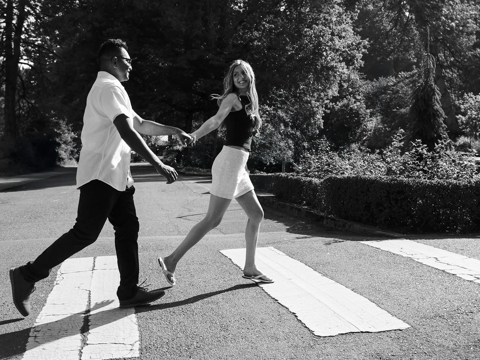Black and white shot of a couple holding hands and crossing a sunlit crosswalk, she glancing back with a smile.