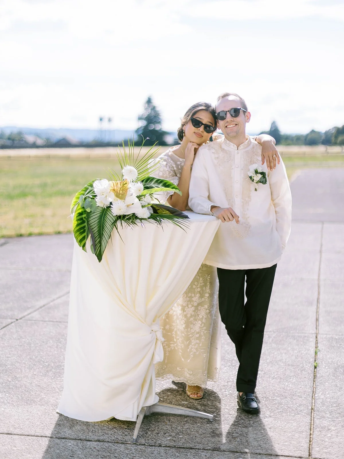 Bride and groom in sunglasses posing together beside a draped cocktail table decorated with a tropical floral arrangement on the airfield tarmac
